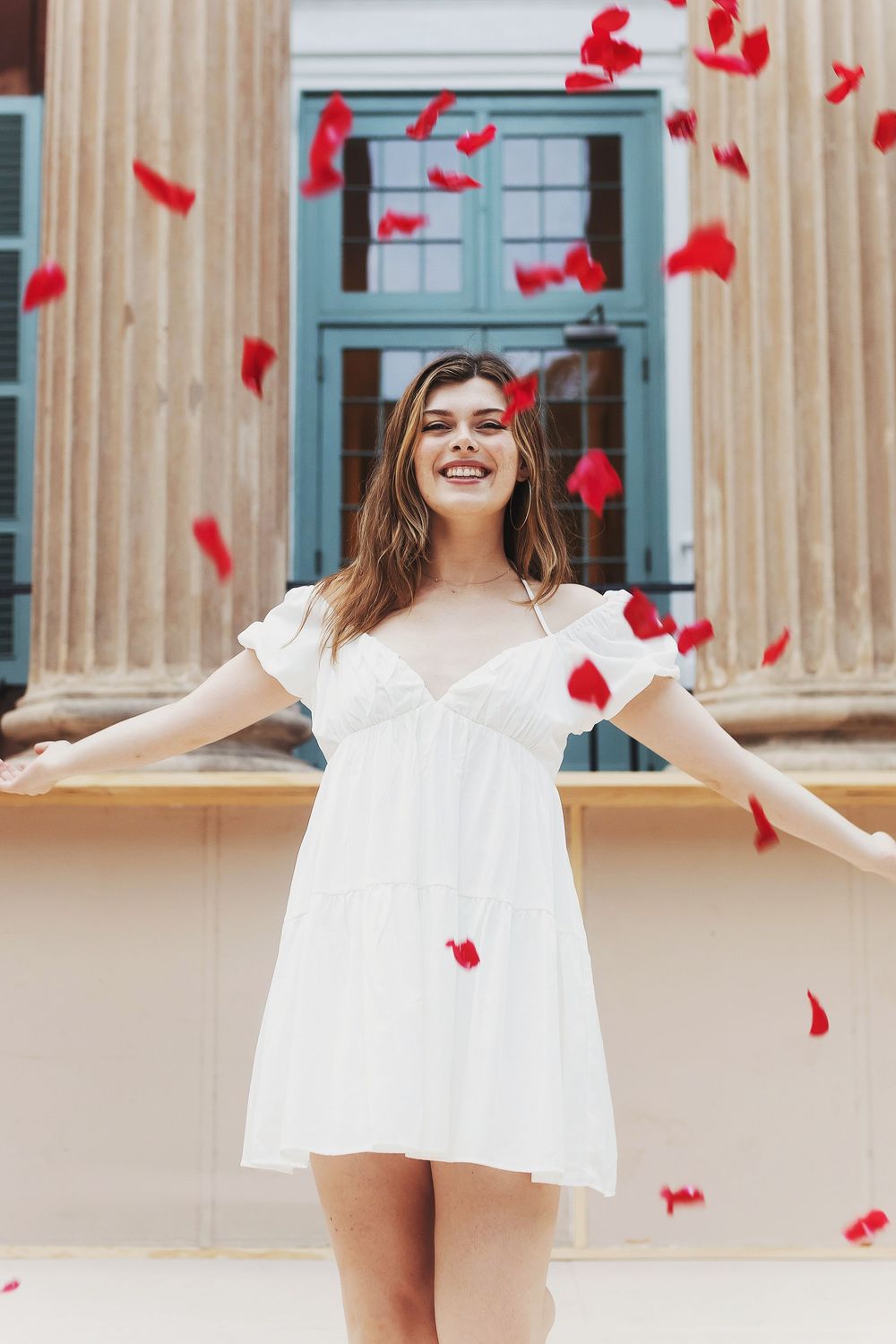 White dress photoshoot with red rose petals falling against a classical building with columns.