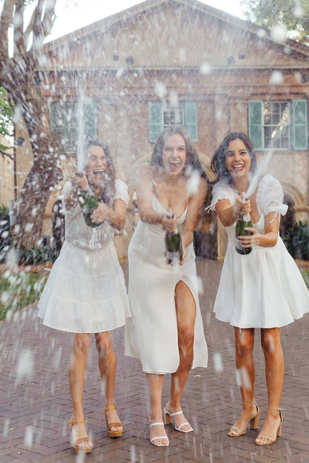 Three friends in white dresses joyfully toss confetti while celebrating outdoors in a courtyard.