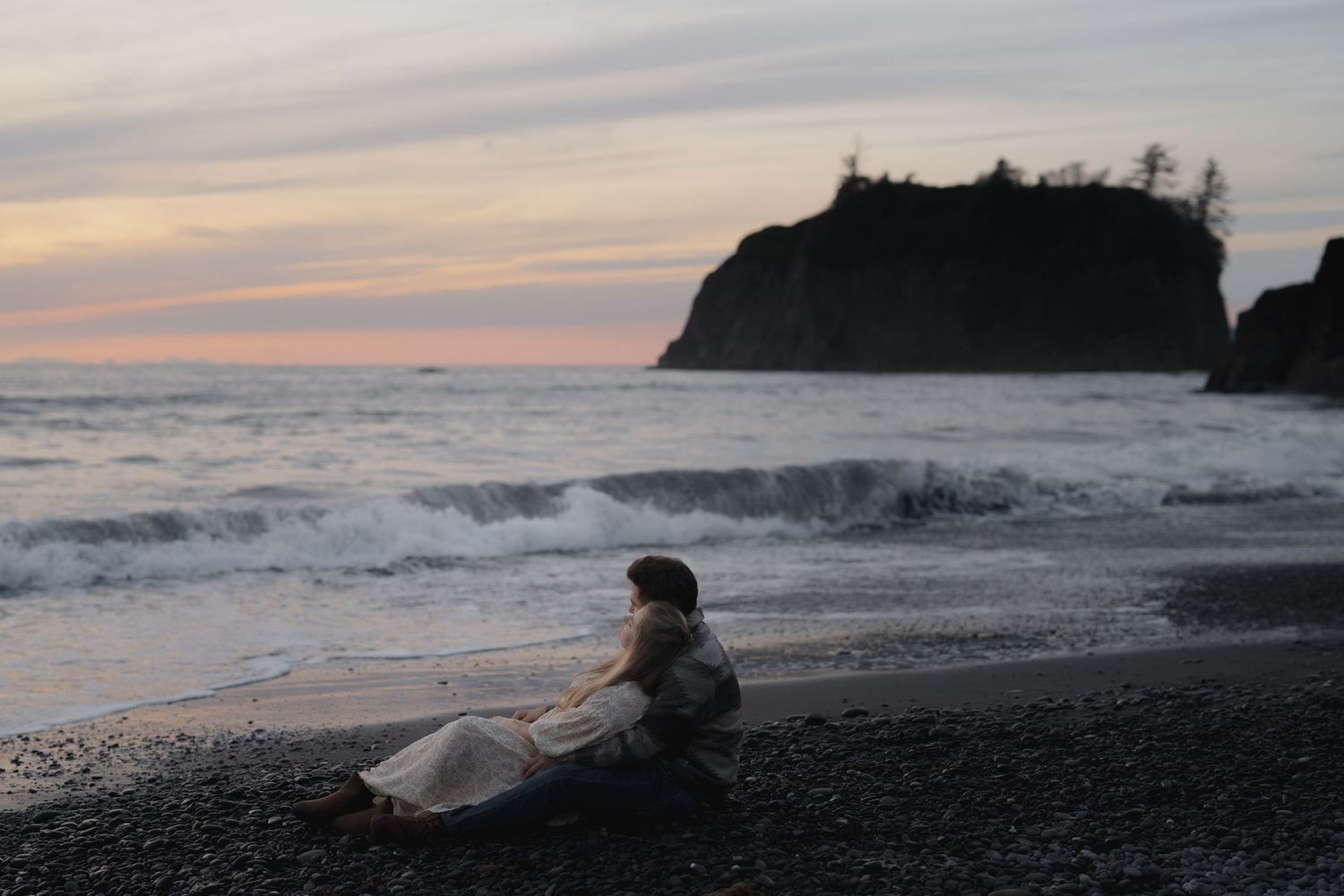 Figure sits alone on dark beach at sunset with dramatic cliffs and crashing waves in the background.