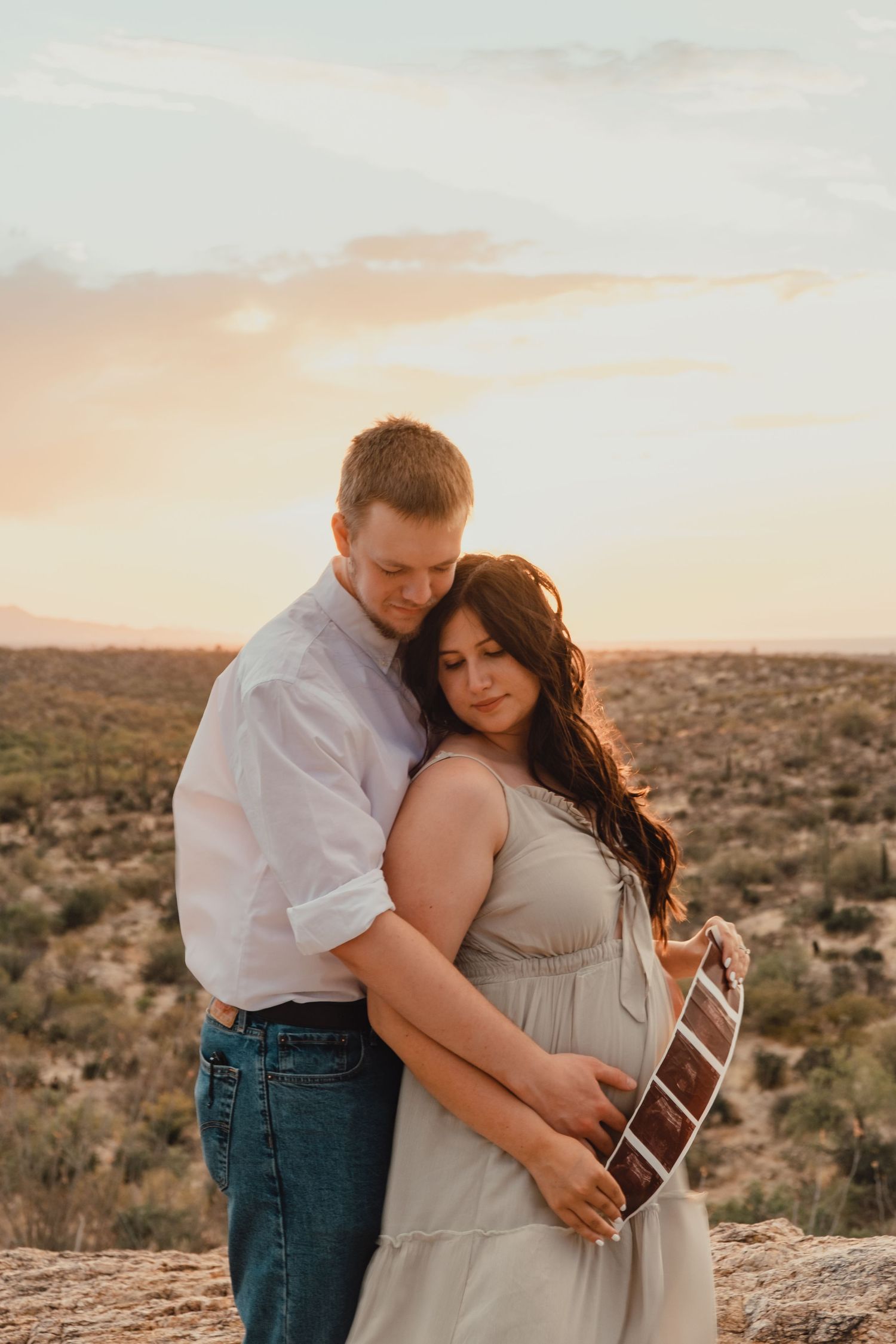 A couple embraces in a desert landscape during golden hour sunset, with desert brush and mountains in the background.