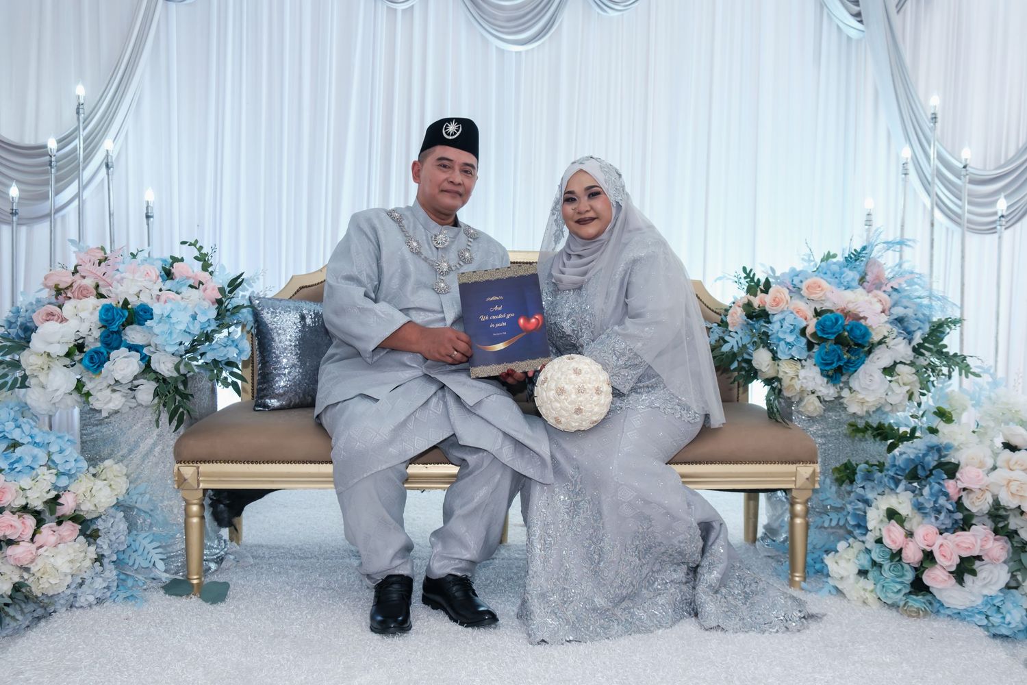 Couple in traditional wedding attire seated on bench with blue floral decorations.