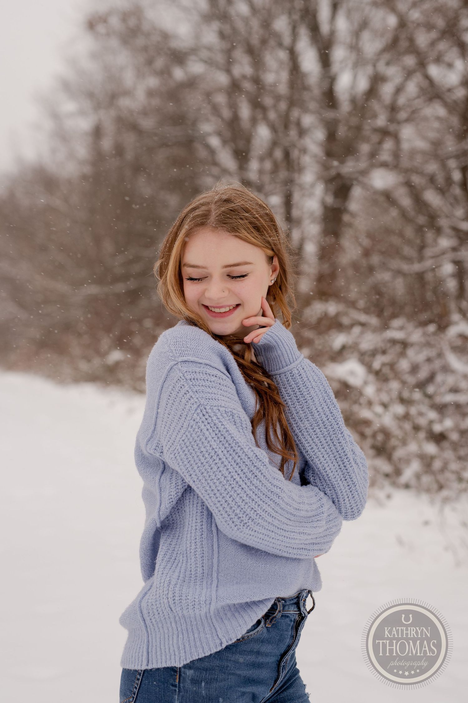 A winter portrait of someone in a cozy blue sweater smiling with snow-covered trees in the background.