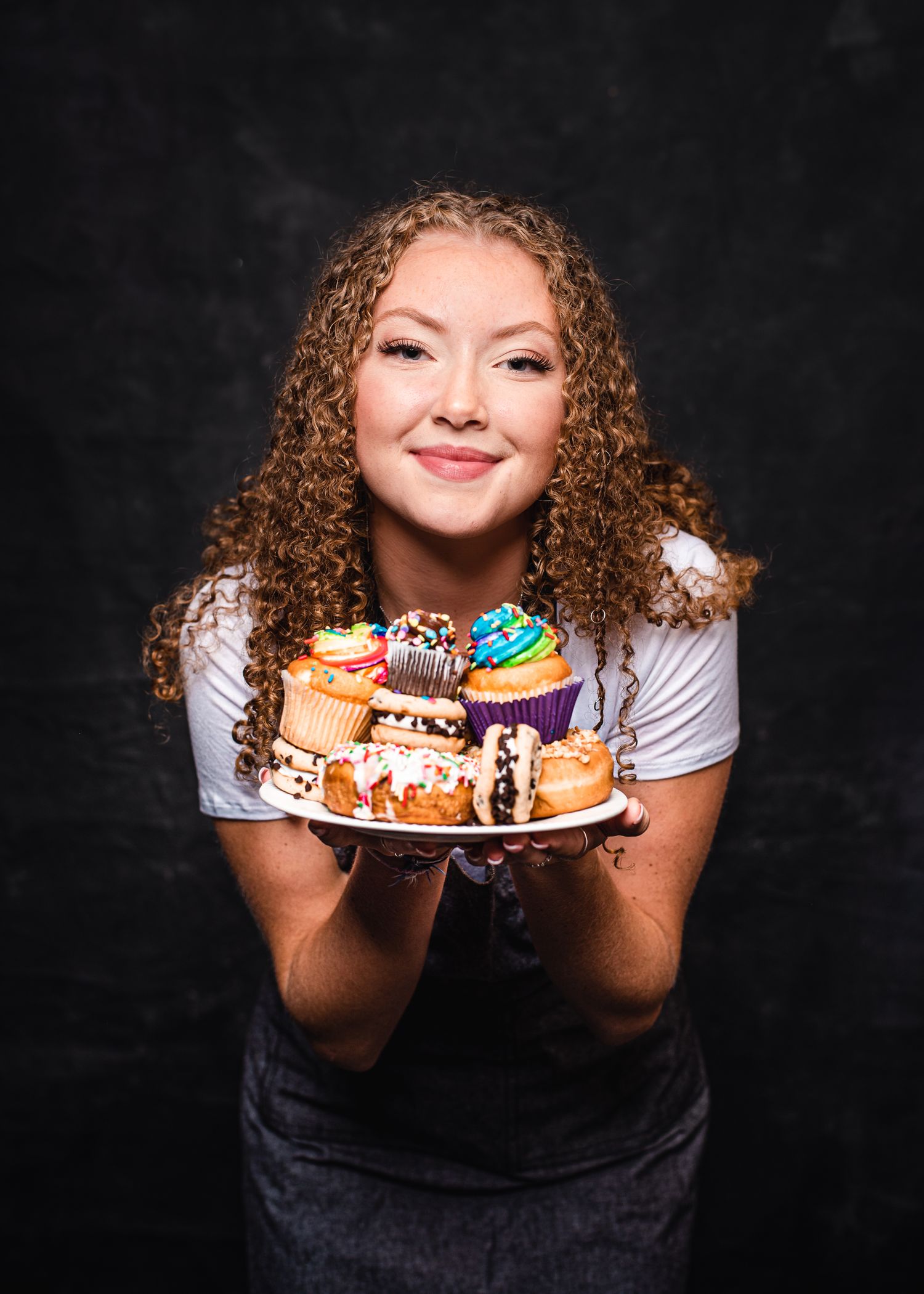 Portrait of curly-haired person holding plate of colorful cupcakes against dark background.