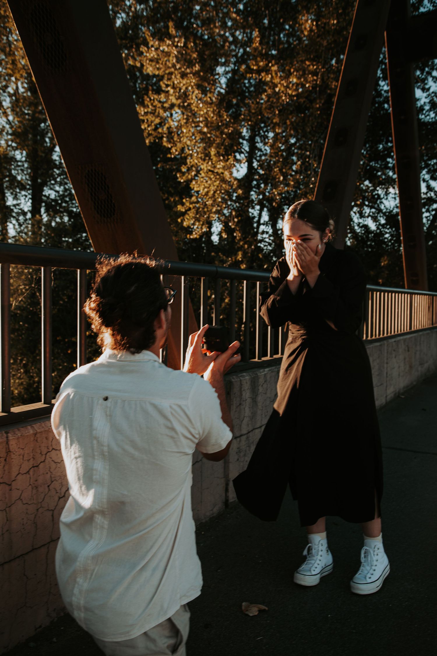 A man in a white shirt kneels with a camera as a woman in black covers her mouth in surprise on a bridge.