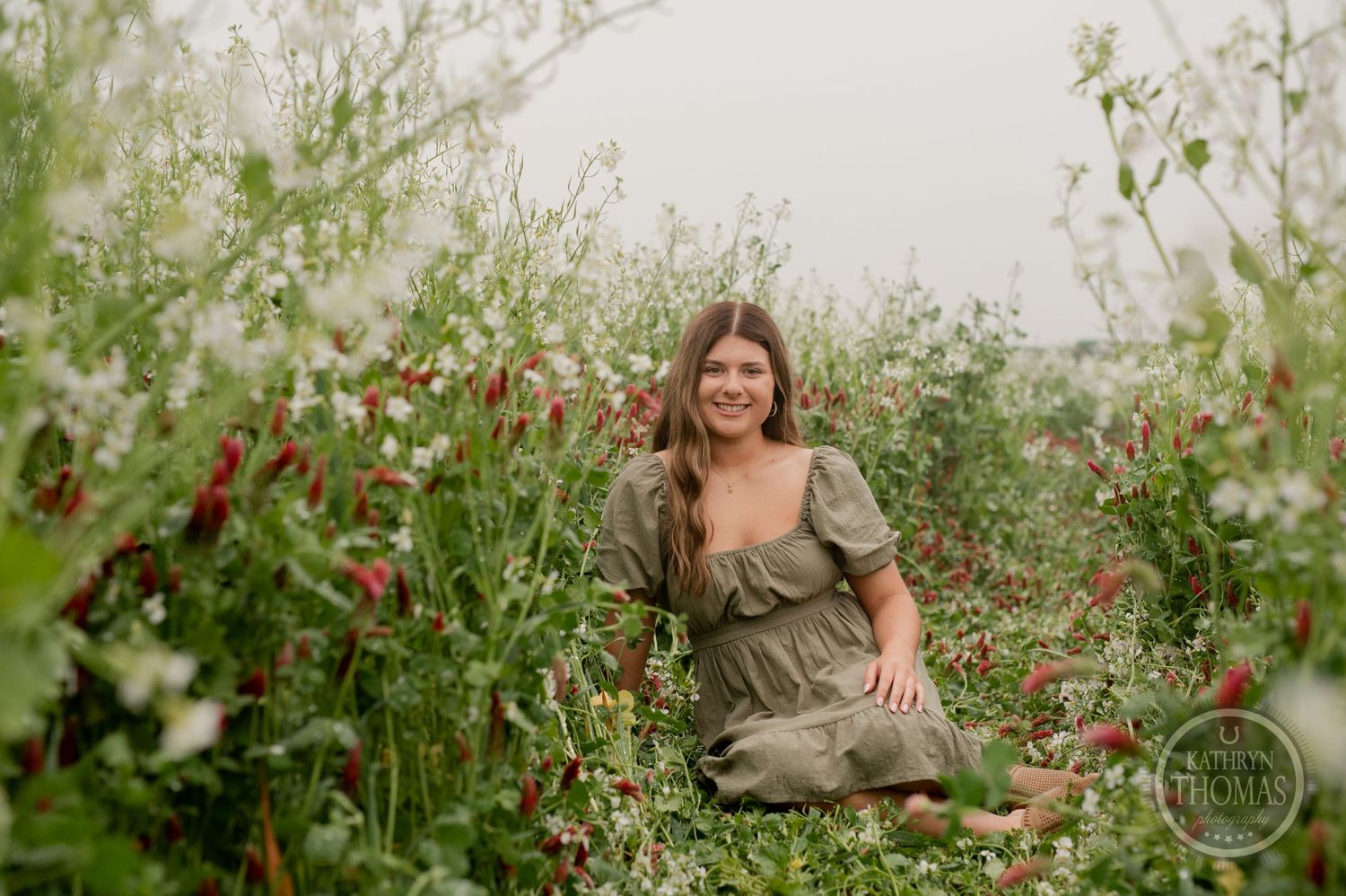 Model sits among blooming wildflowers with vintage chair in rustic outdoor setting.