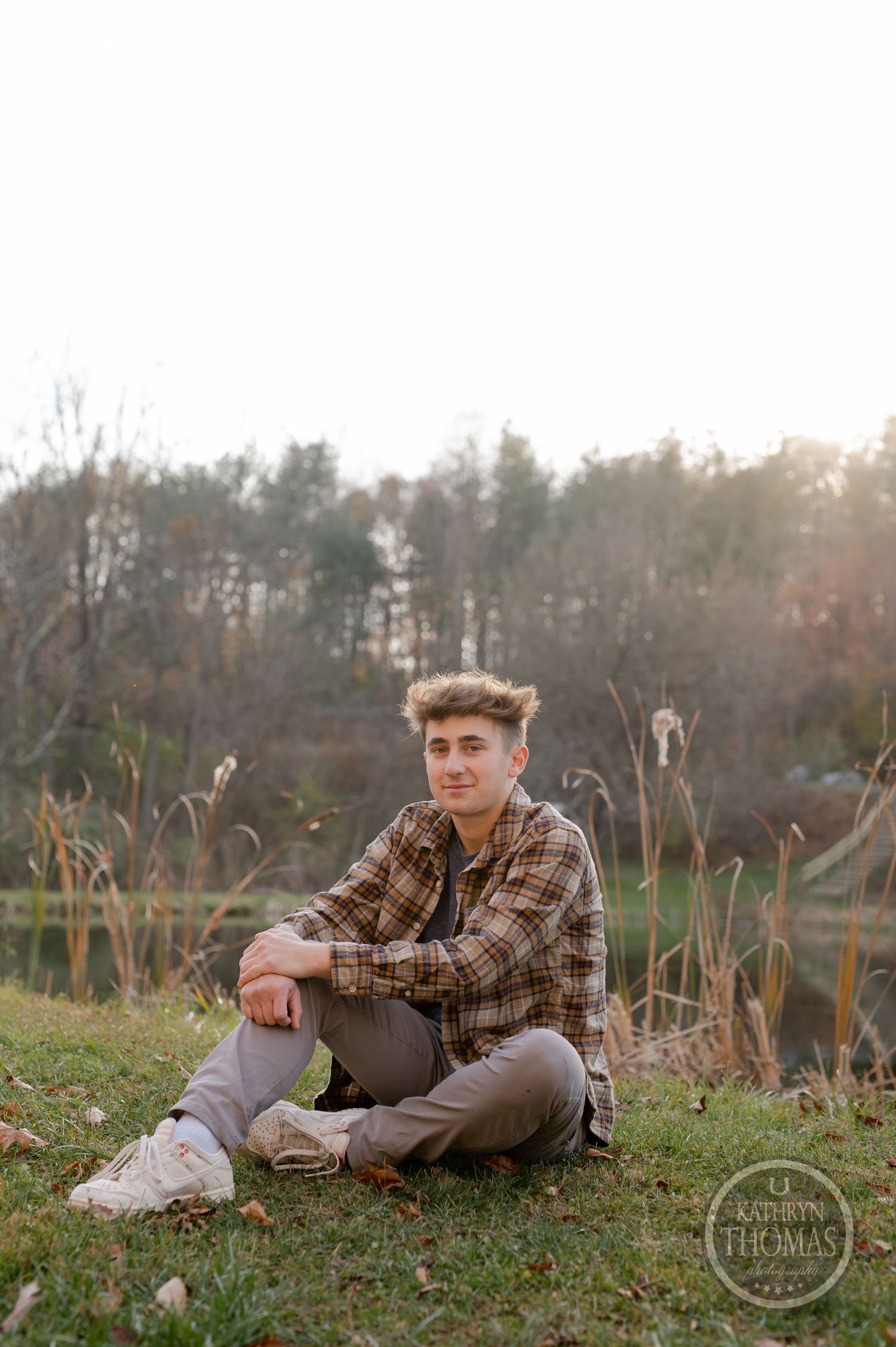 A person sits casually on grass near a lake wearing a brown plaid shirt and khaki pants.