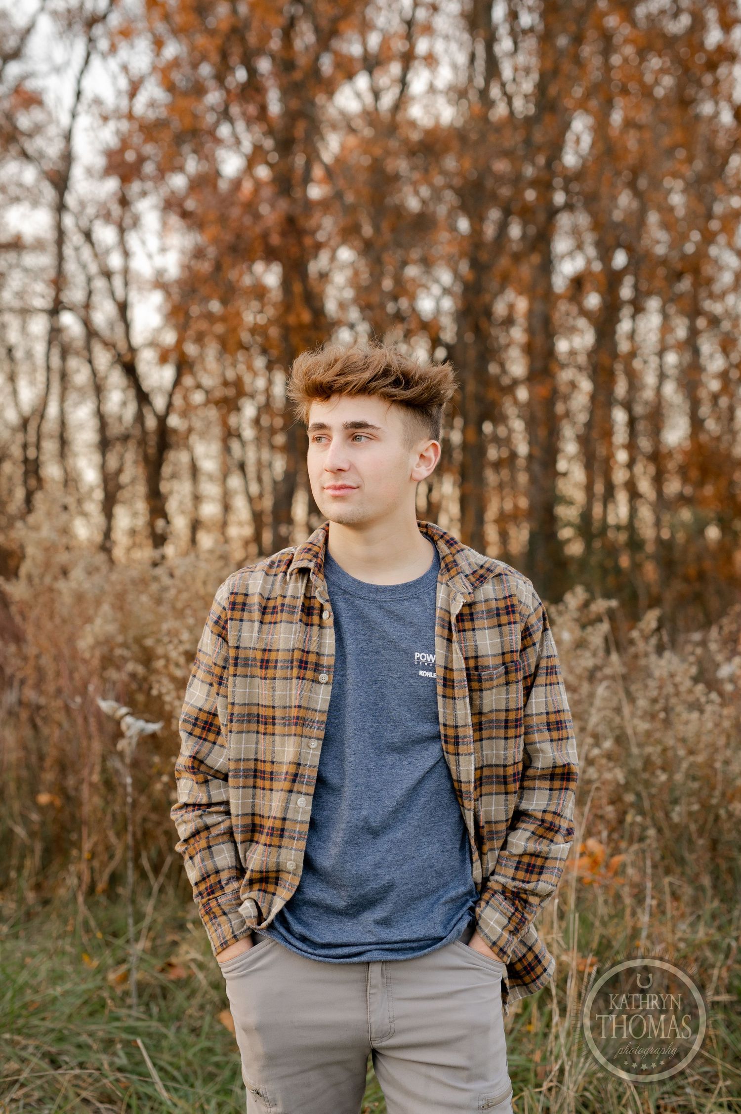 Someone in a plaid button-down shirt and blue t-shirt stands in an autumn forest setting.