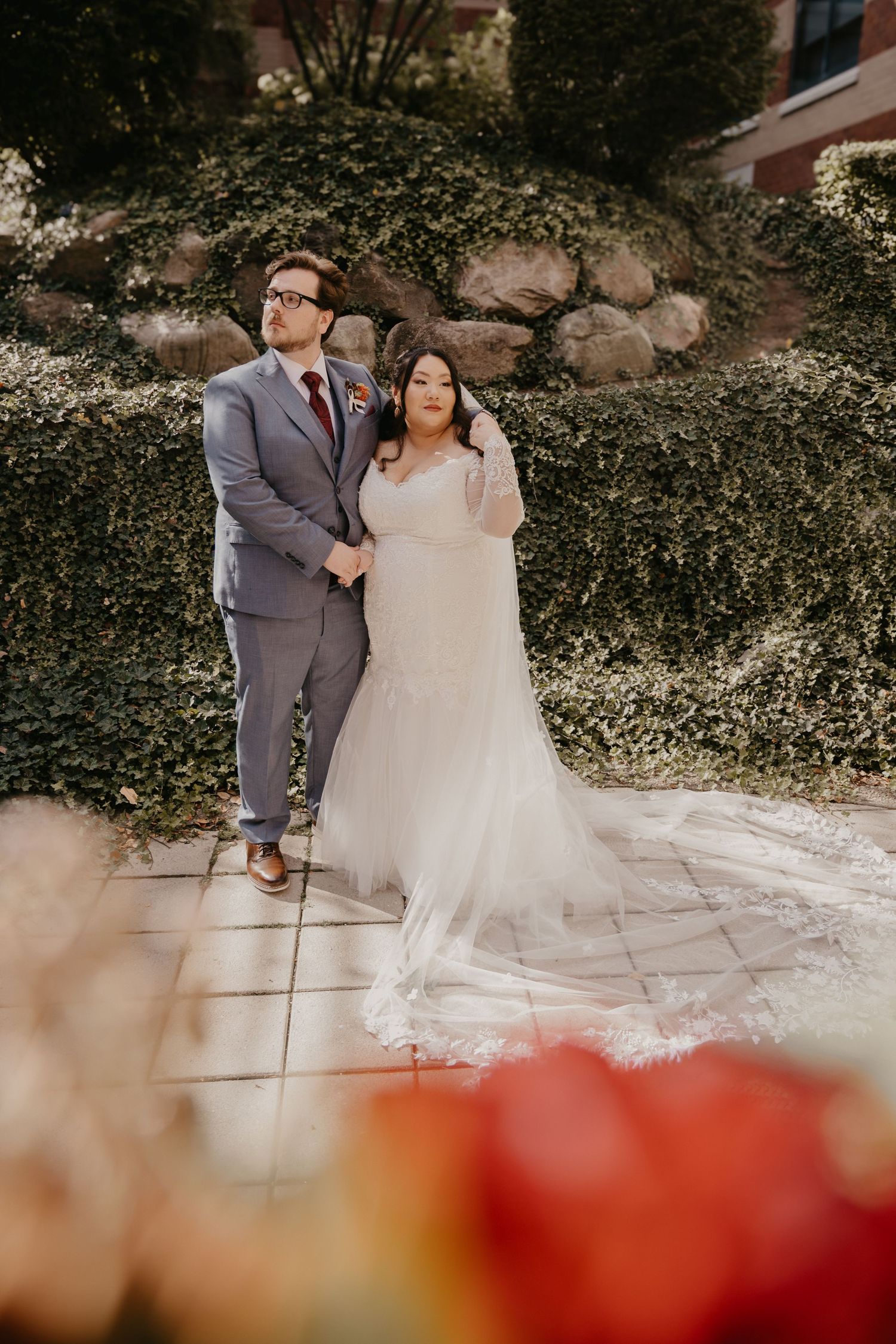 Bride in flowing white gown and groom in gray suit posing before lush green hedge wall.