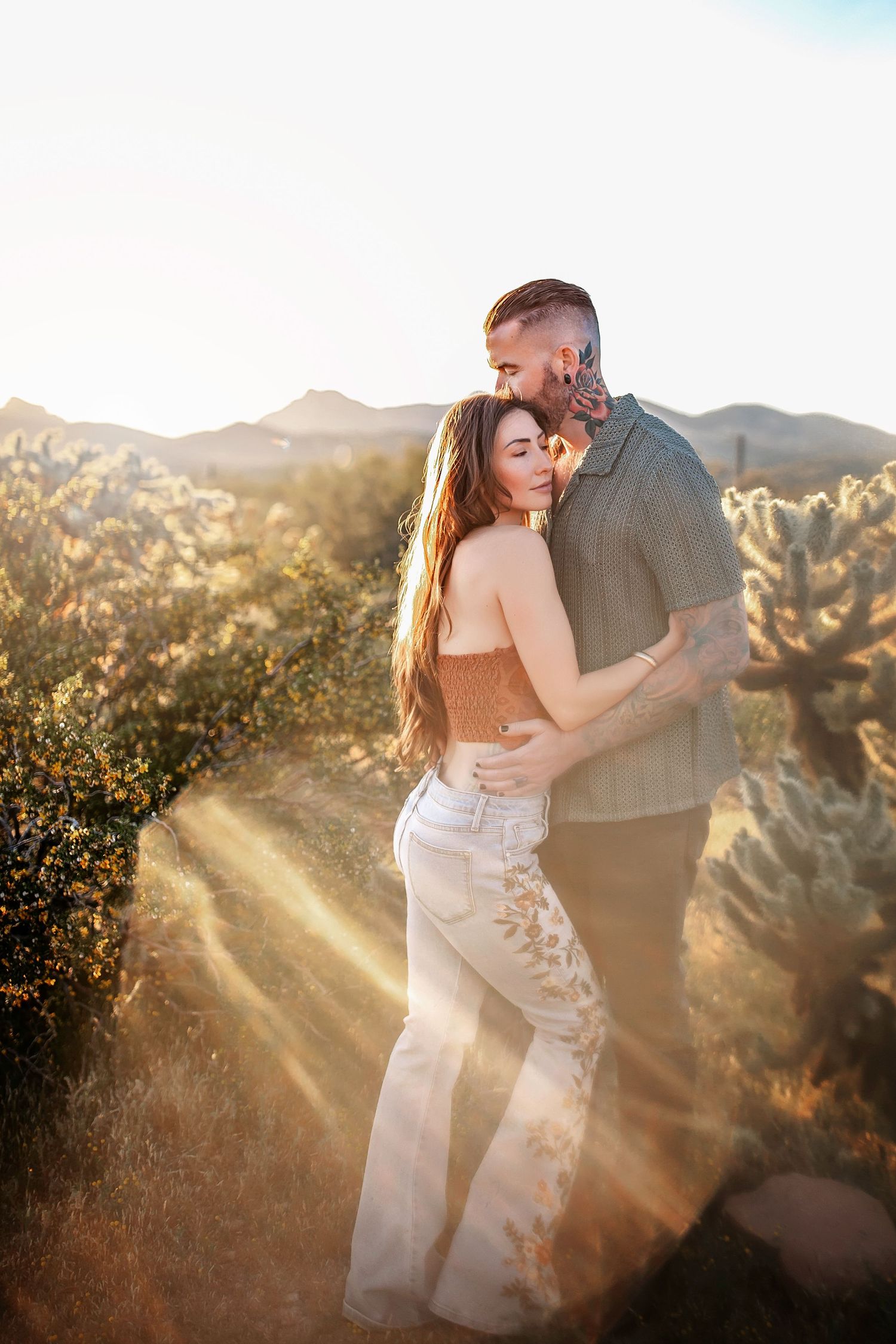 Couple embraces in desert setting with golden sunlight streaming through cacti.