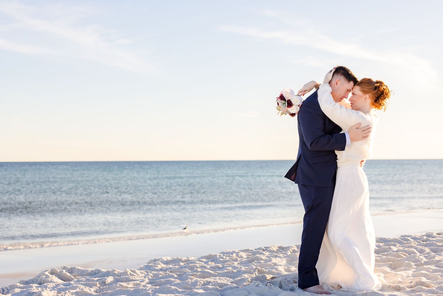 A romantic beach wedding couple dances together on white sand with the ocean and blue sky in the background.
