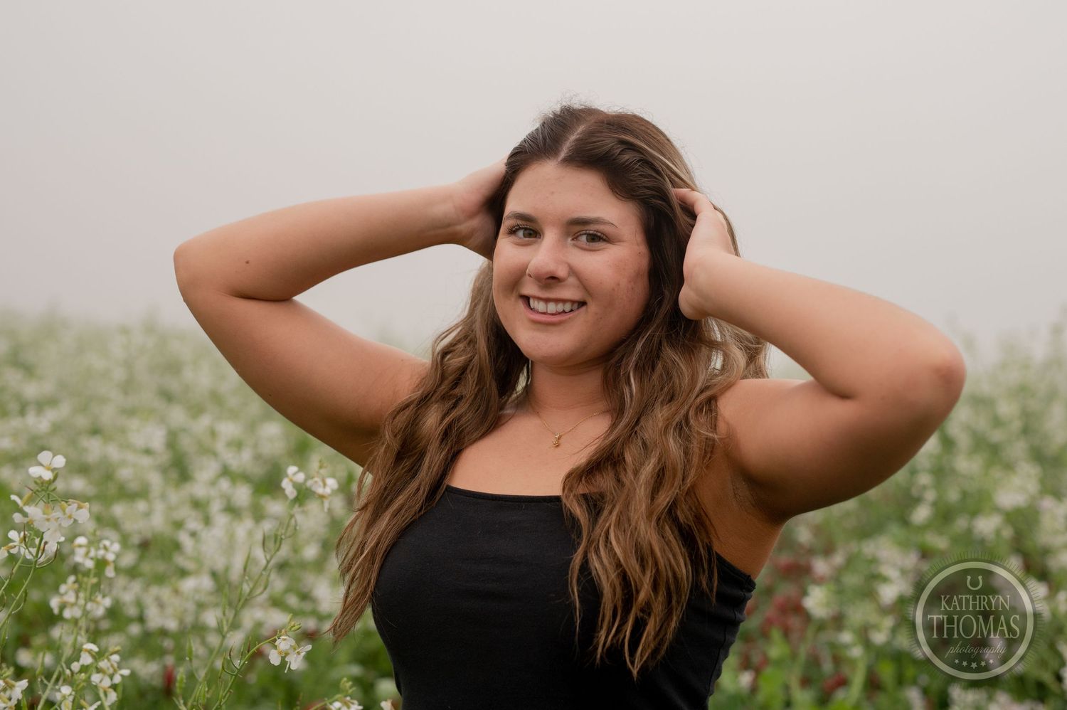 Natural outdoor portrait with hands in hair surrounded by blooming wildflowers on an overcast day.