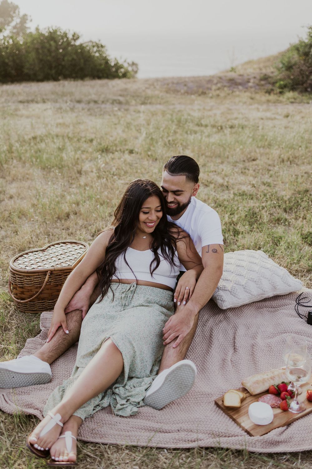 Couple relaxes on picnic blanket with basket and snacks in rustic outdoor setting.