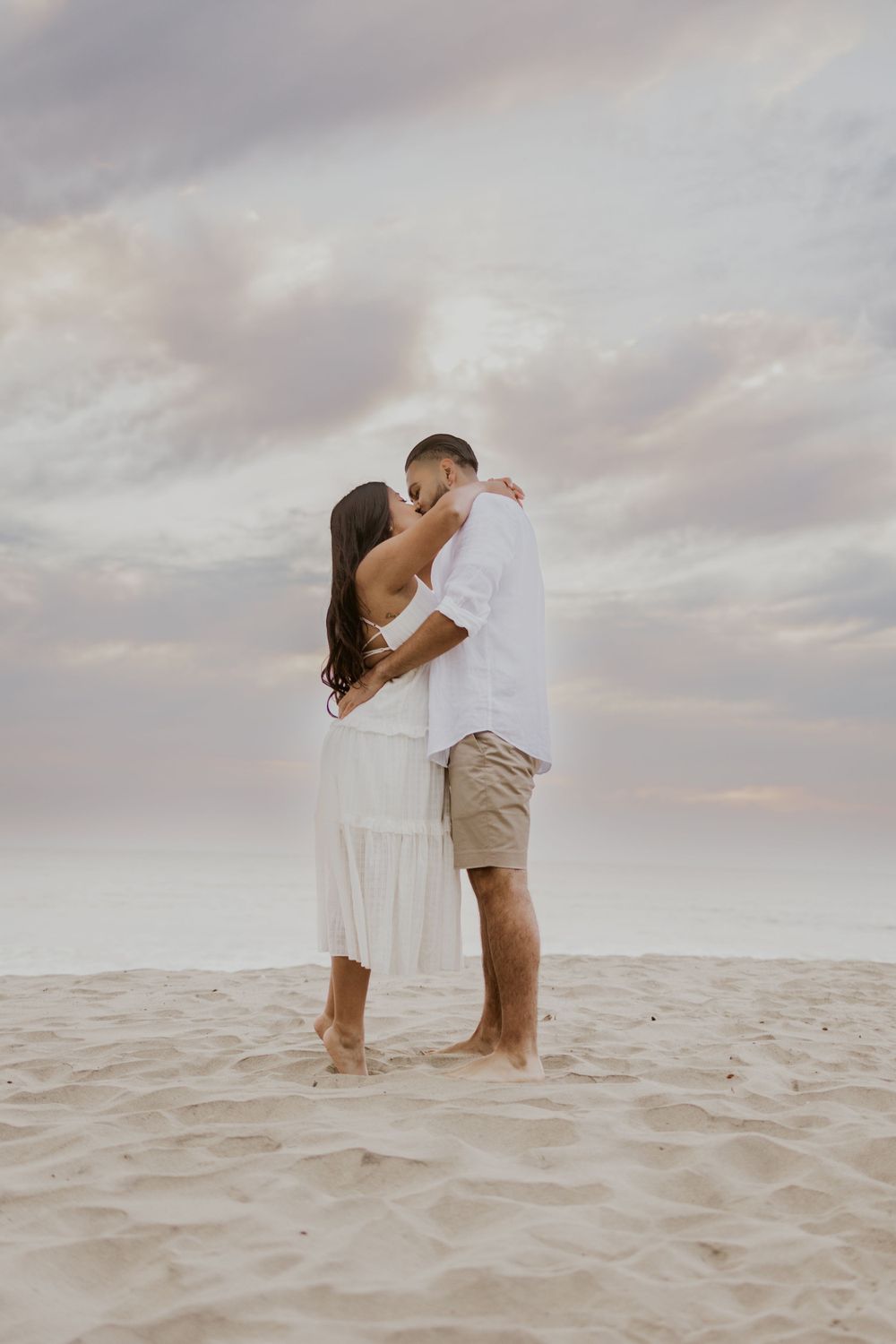 Romantic couple embraces on sandy beach during sunset with cloudy pink sky.