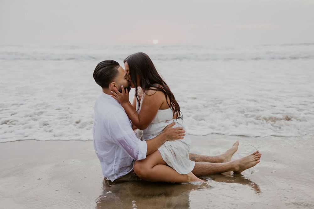 Couple sharing an intimate moment while sitting on wet sand at the beach during sunset.