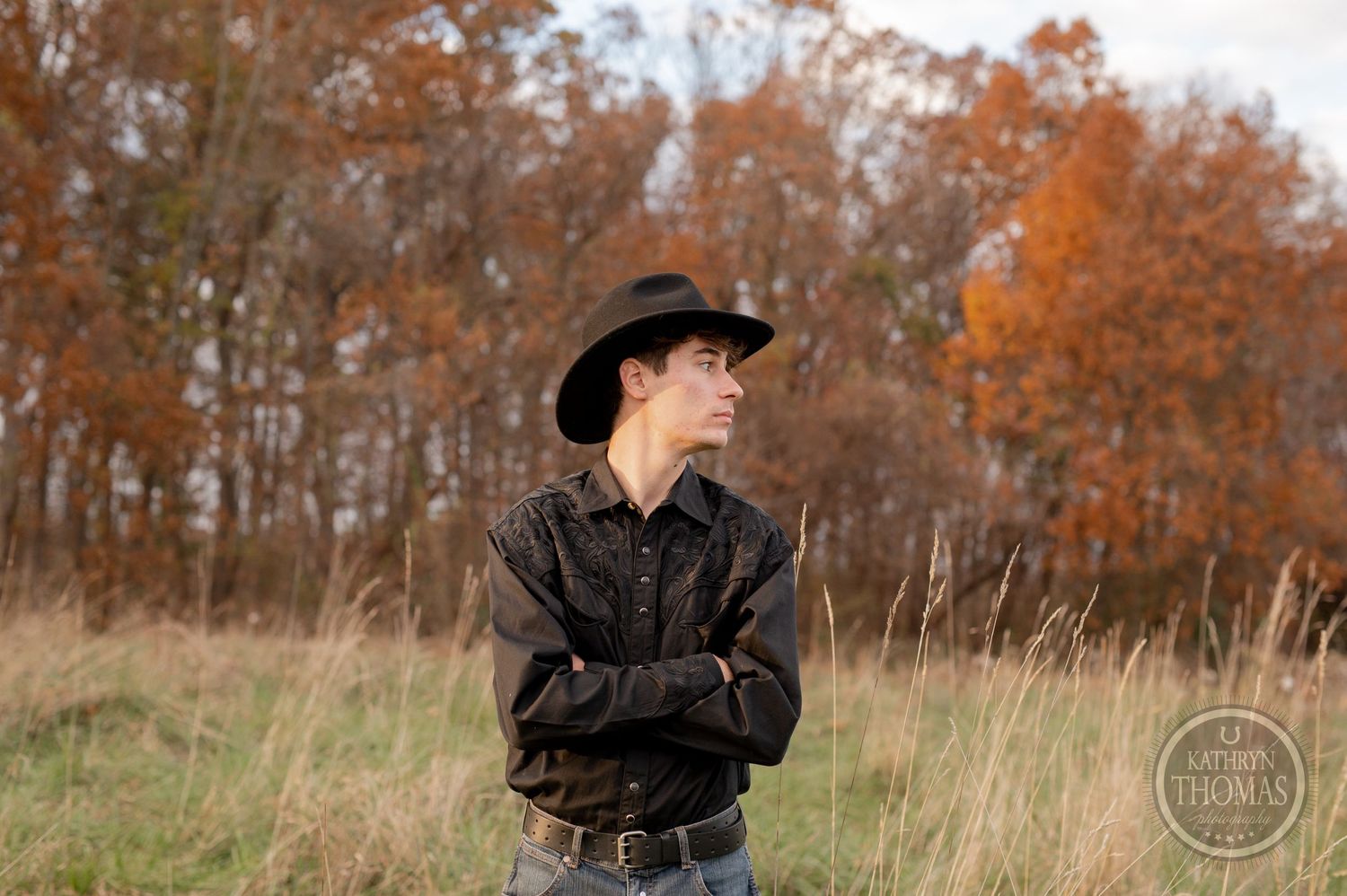 Profile portrait with crossed arms against autumn trees and tall grass.