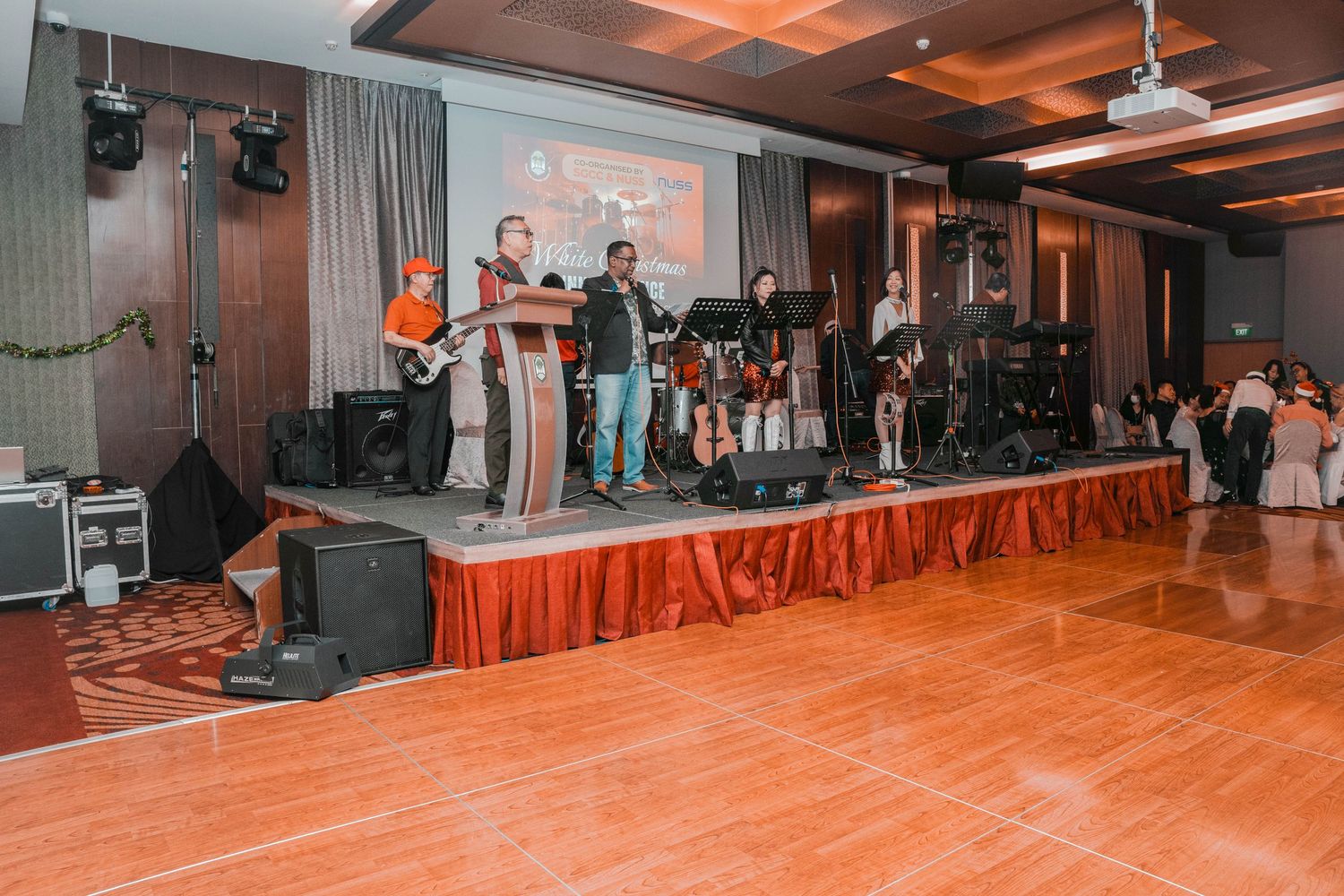 Live band performing on a raised stage with orange curtain draping and wooden dance floor in an indoor venue.