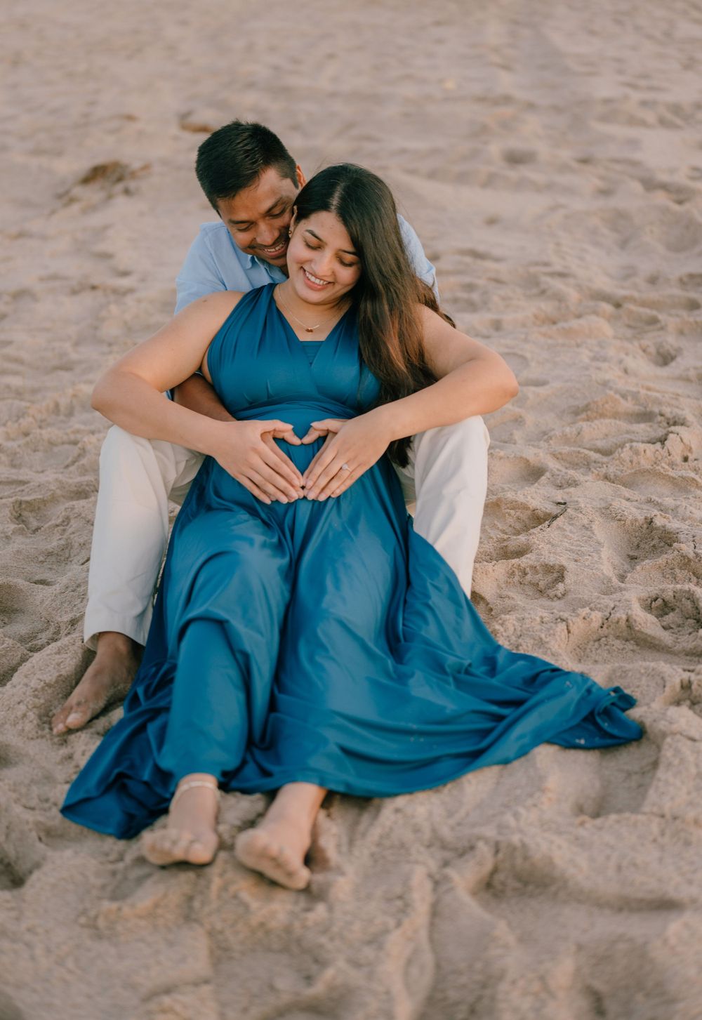 A couple sharing an intimate moment on the beach while making a heart shape over a baby bump.