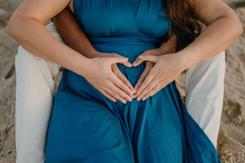 Hands forming a heart shape over a pregnant belly in a blue dress on the beach.
