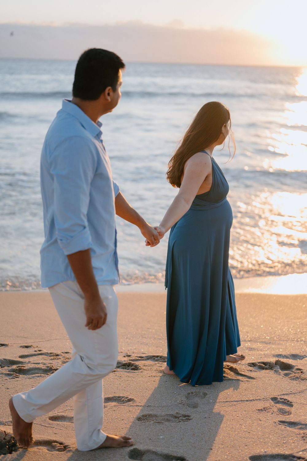 A couple holding hands walks along a beach at sunset with waves crashing in the background.