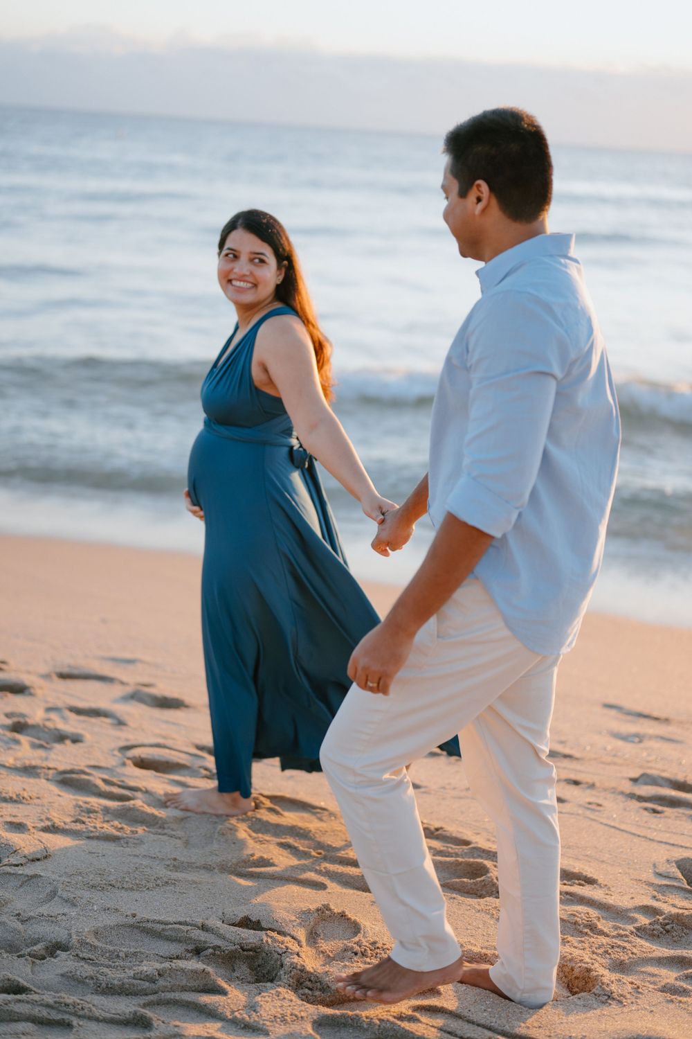 Expectant couple in coordinating blue and white outfits stroll together on a sandy beach.