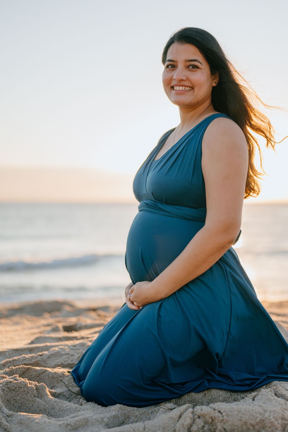 Woman in flowing blue dress poses on beach sand during golden hour.