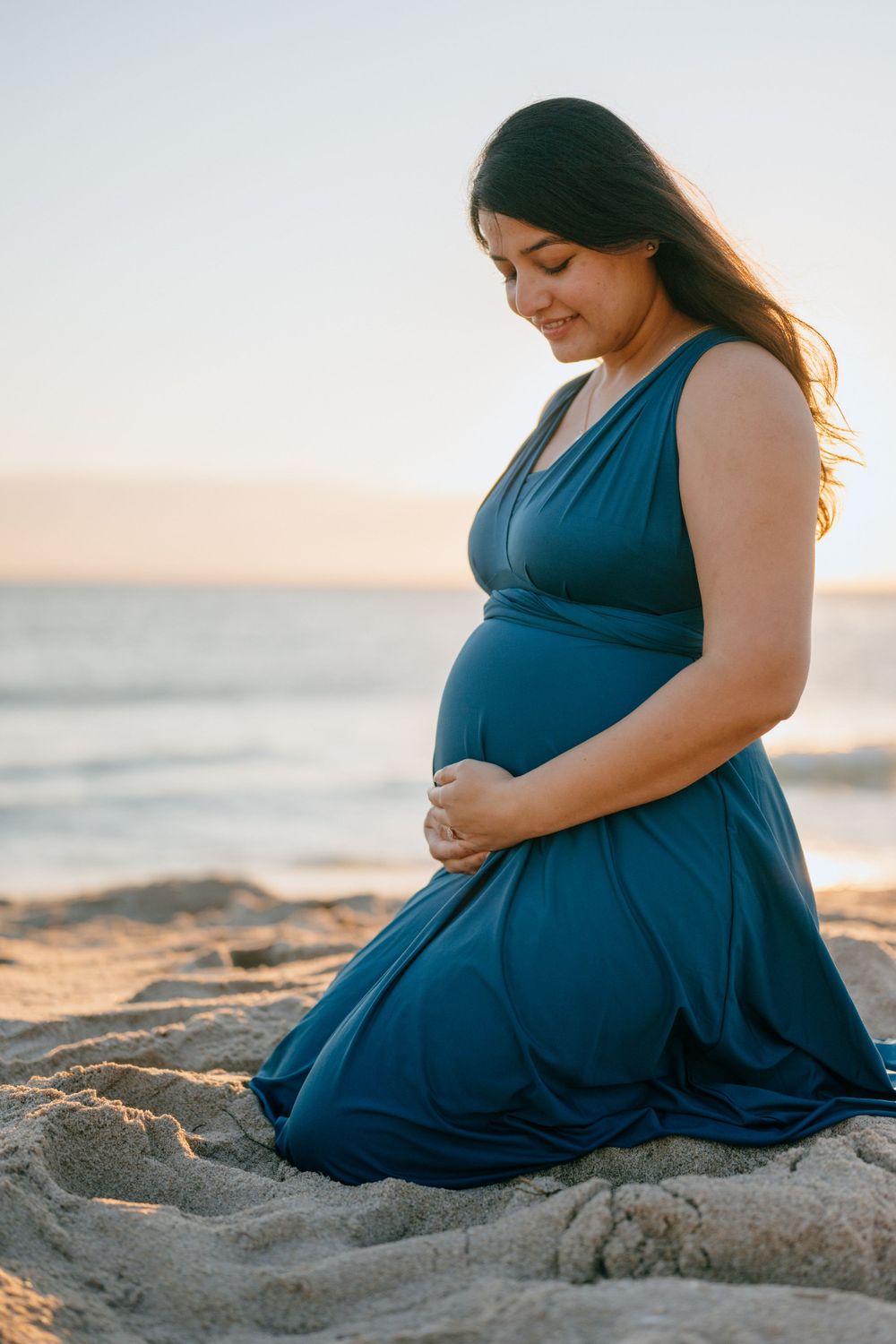Woman in teal dress sits on beach sand at sunset, cradling her baby bump.