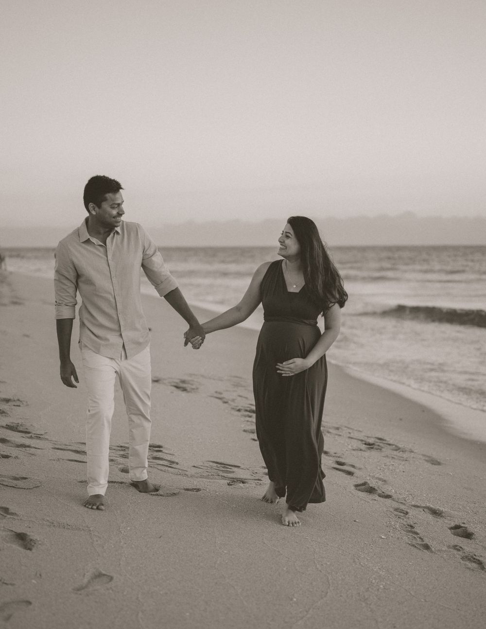 A black and white beach photo of an expecting couple walking hand in hand along the shore.