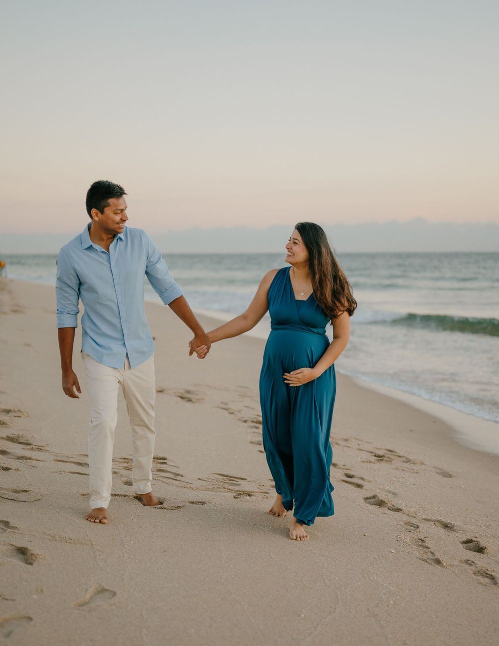 Expectant couple walking hand in hand on beach at sunset.