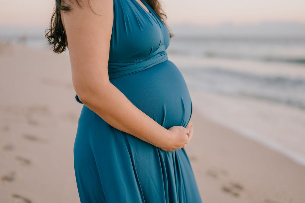 Close-up of a pregnant belly in a teal blue dress on a sandy beach at sunset.