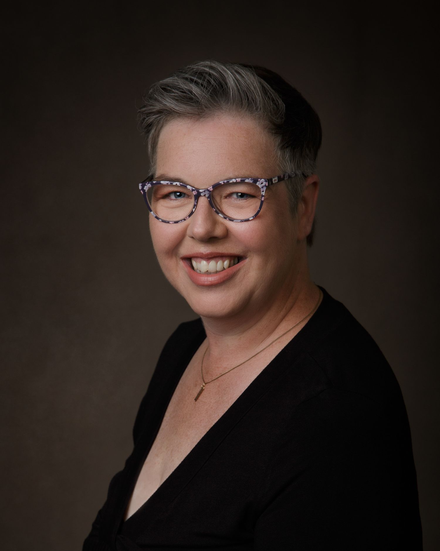 A woman wearing glasses and a black top smiles warmly at the camera.