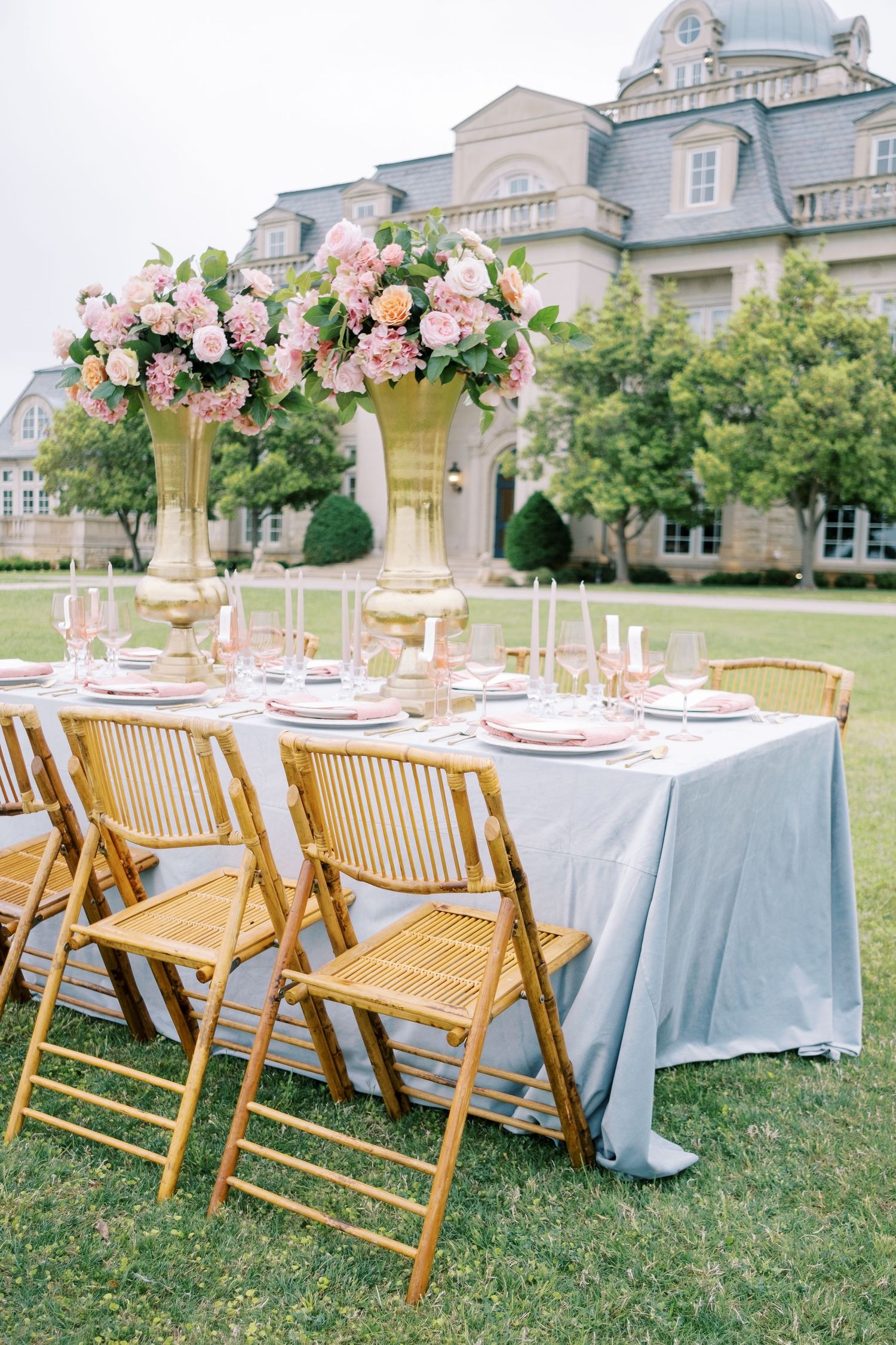 Elegant outdoor wedding table with pink floral centerpiece and gold chairs against mansion backdrop.