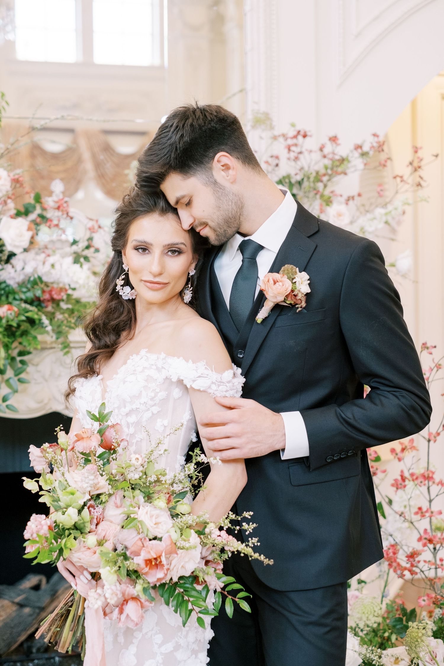 A romantic wedding portrait featuring an elegant off-shoulder lace gown and pink floral bouquet against a light backdrop.
