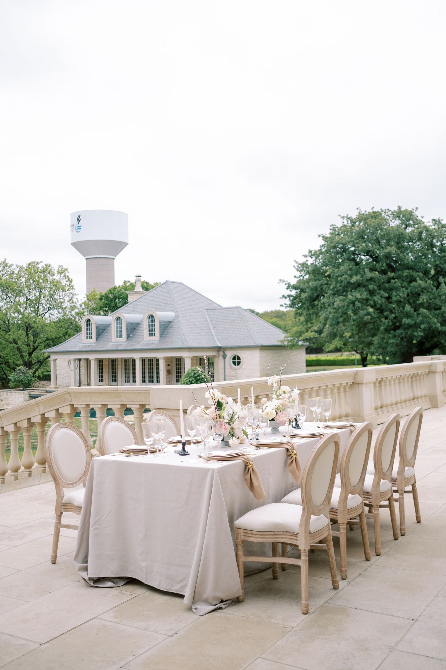 An outdoor wedding reception table setup with white linens and French country chairs on a stone terrace.