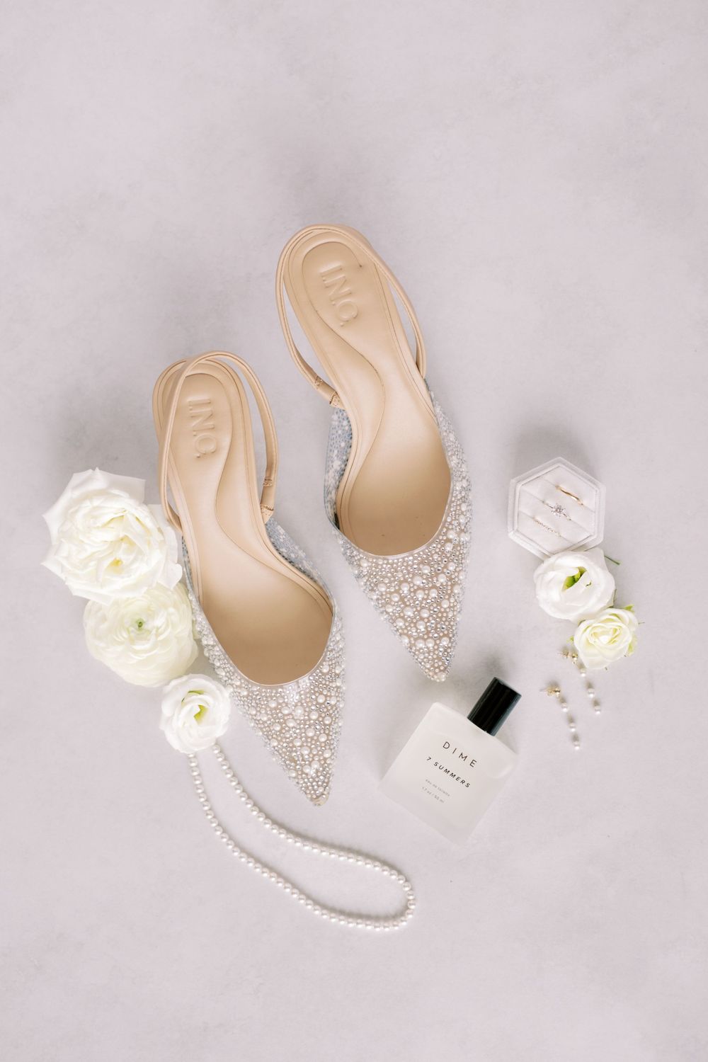 Bridal accessories flatlay with embellished shoes, white roses, and perfume bottle on light gray background.