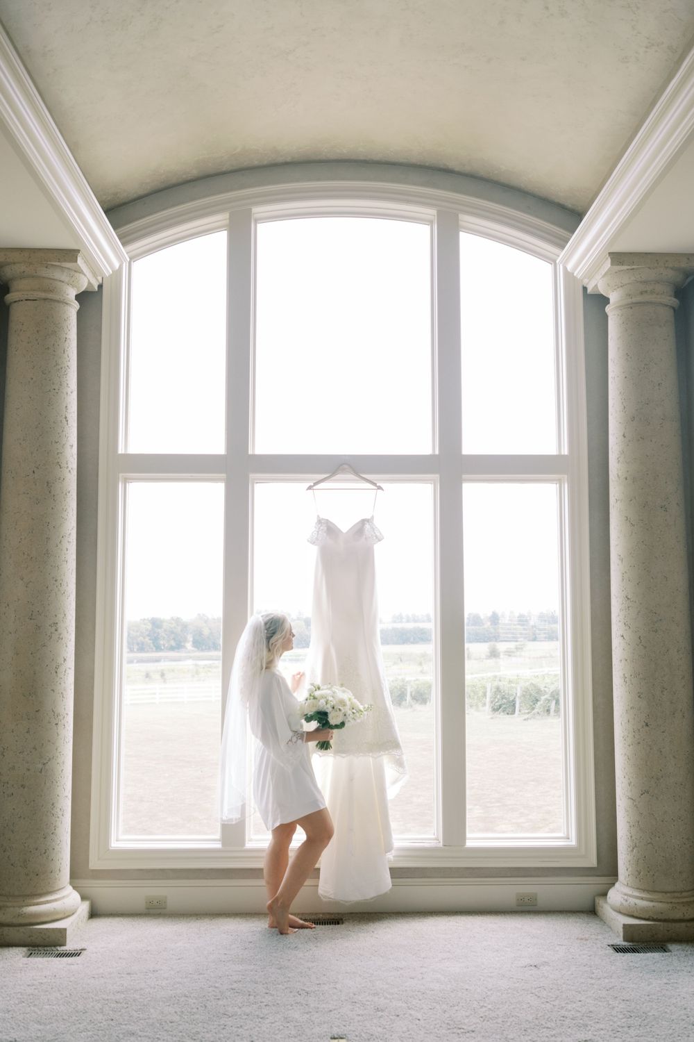 Elegant bridal gown silhouettes photographed against large arched windows in bright interior space.