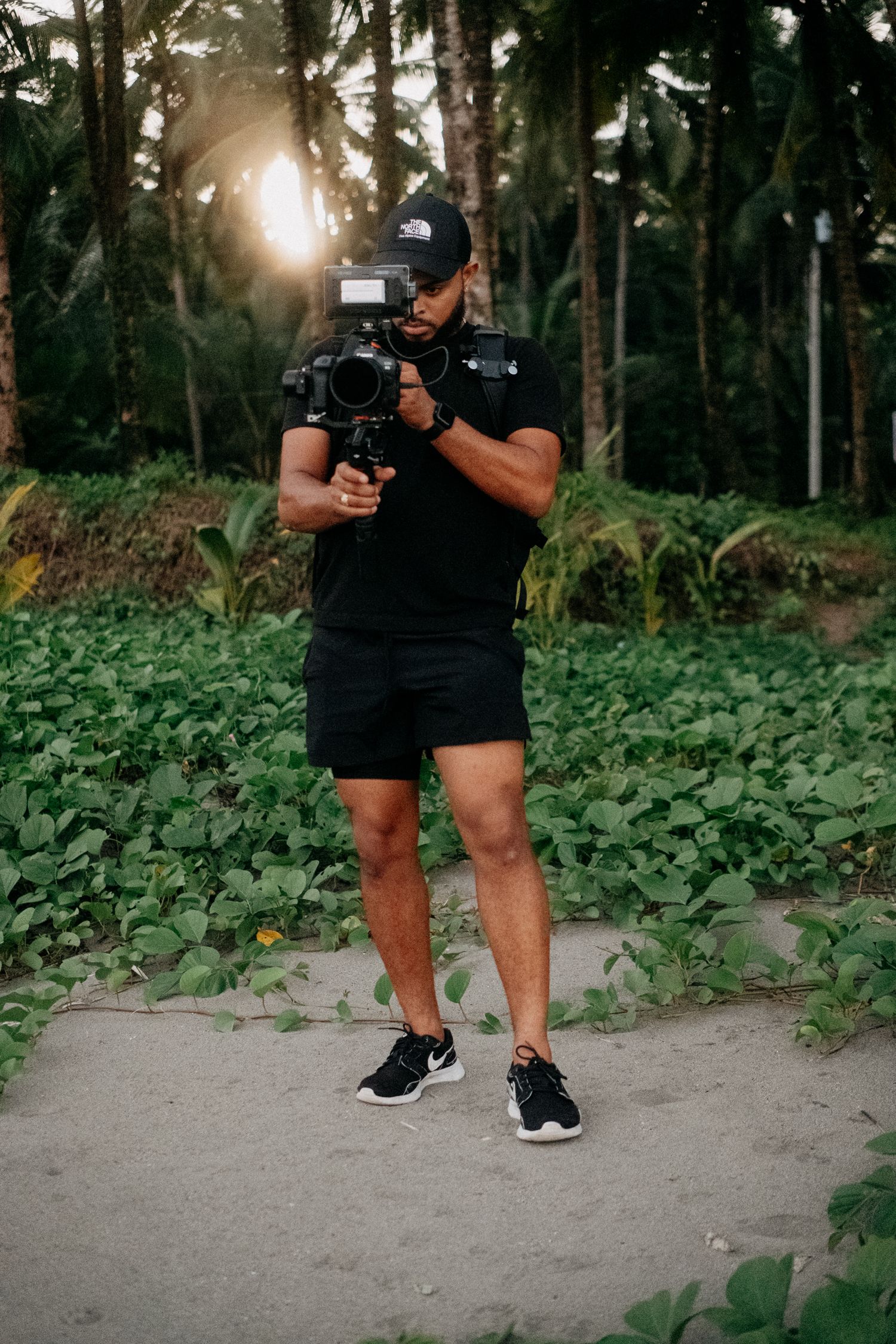 Photographer with camera equipment walking along a tropical path surrounded by lush vegetation.