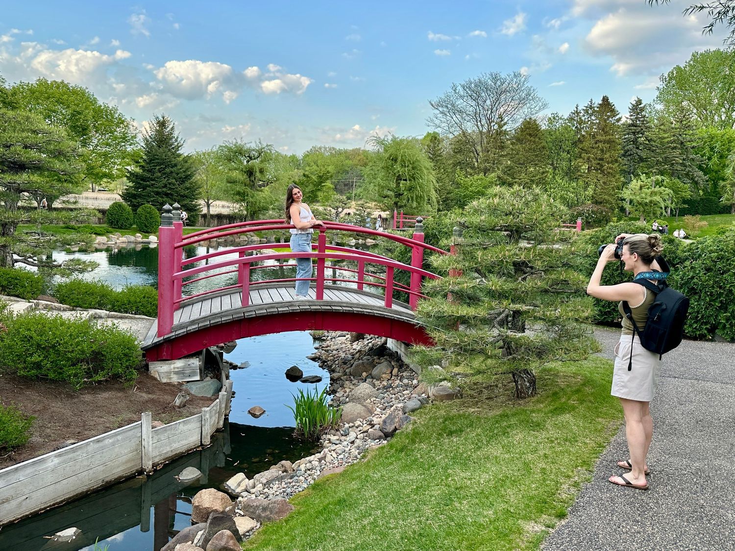 A vibrant red wooden bridge spans a stream in a peaceful Japanese garden while someone photographs the scenic view.