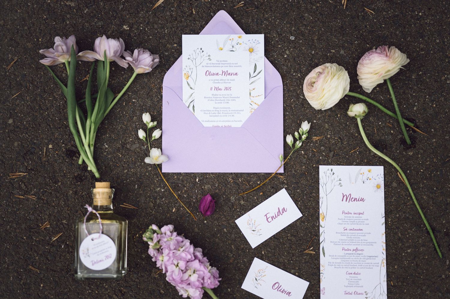 Purple wedding invitation suite with floral details arranged on dark surface with fresh flowers and small glass bottle.