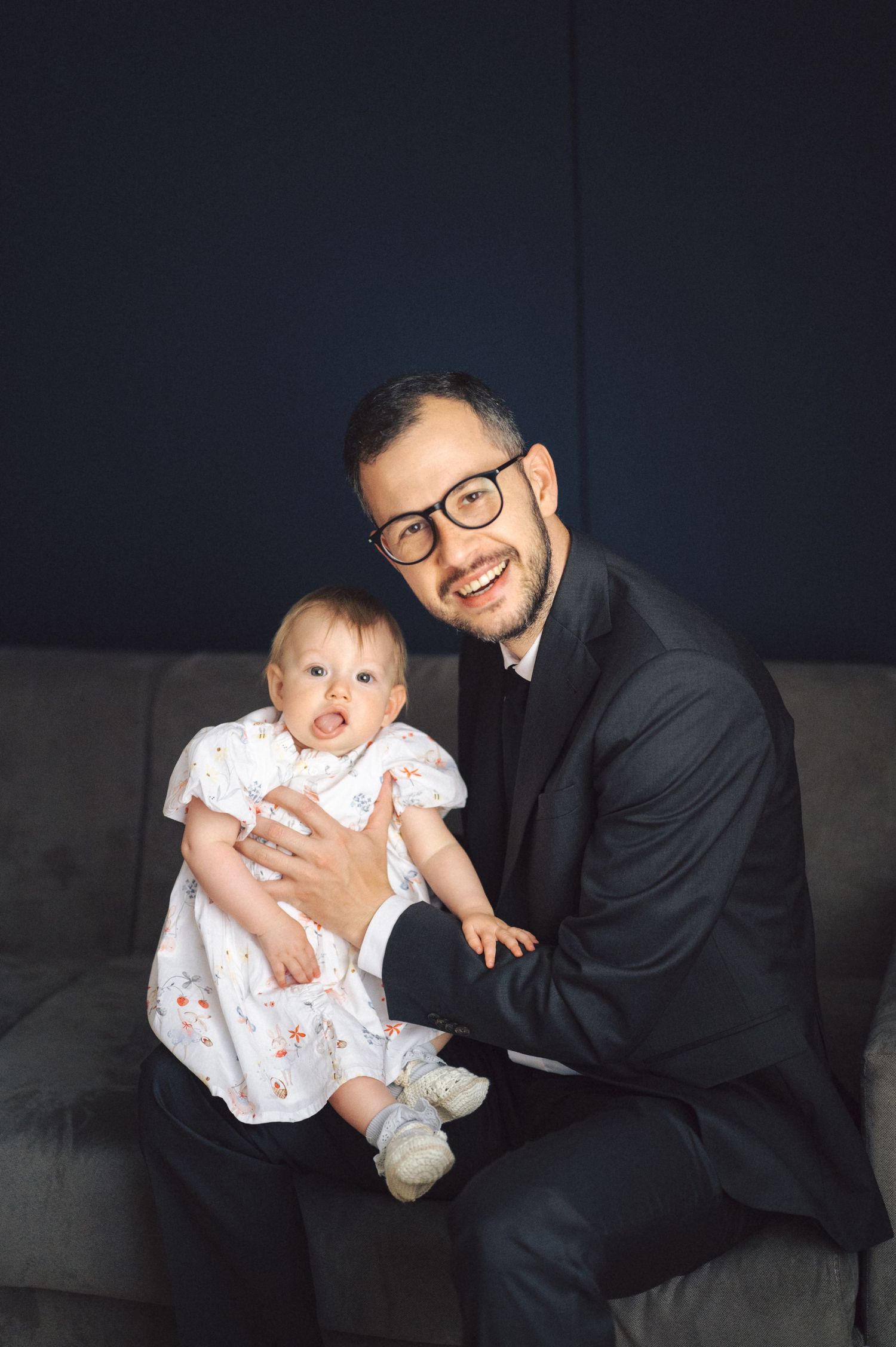 A person in a black suit sits on a gray couch holding a baby in a white floral dress.