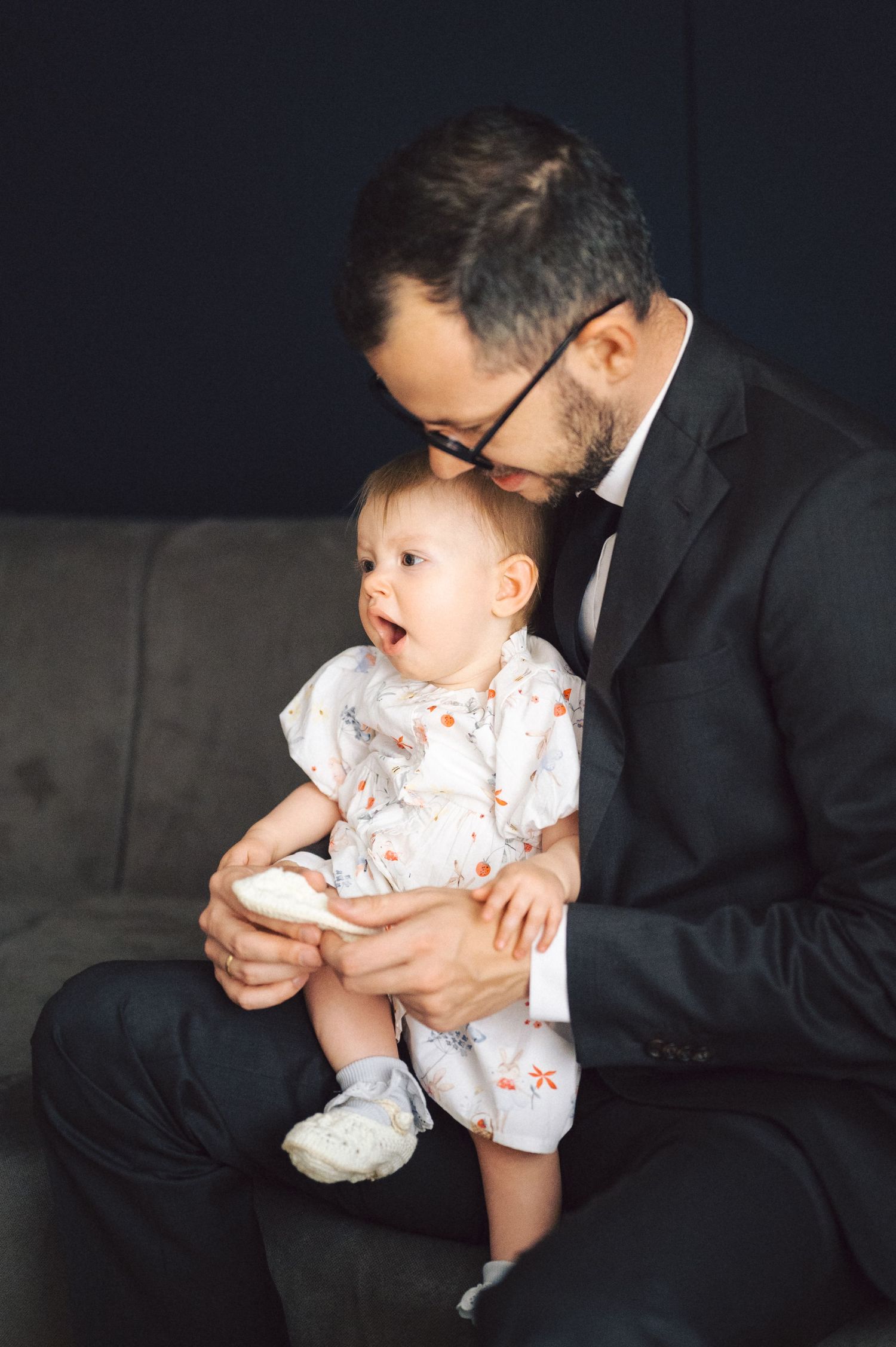 A tender moment between a formally dressed man in a suit and a baby in a white floral outfit sitting together on a couch.