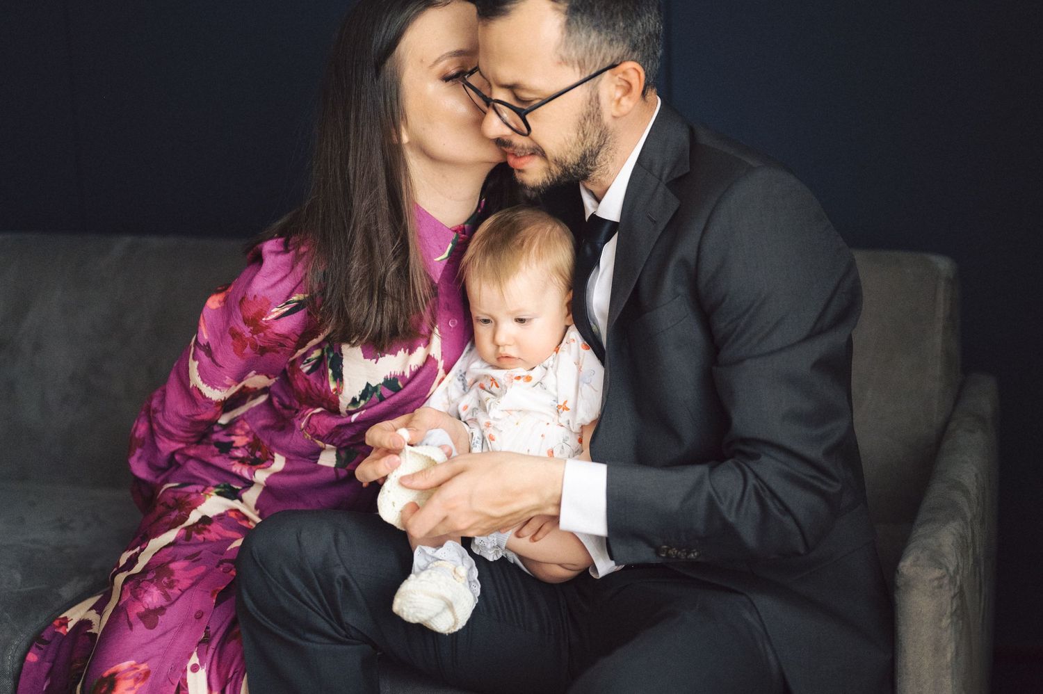 A family sits together on a dark couch, with a parent in an elegant magenta robe and another in a suit holding a baby.