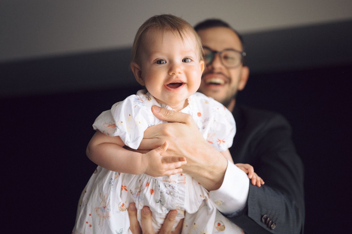 A happy baby wearing a white dress smiles while being held by someone in a black suit and glasses.