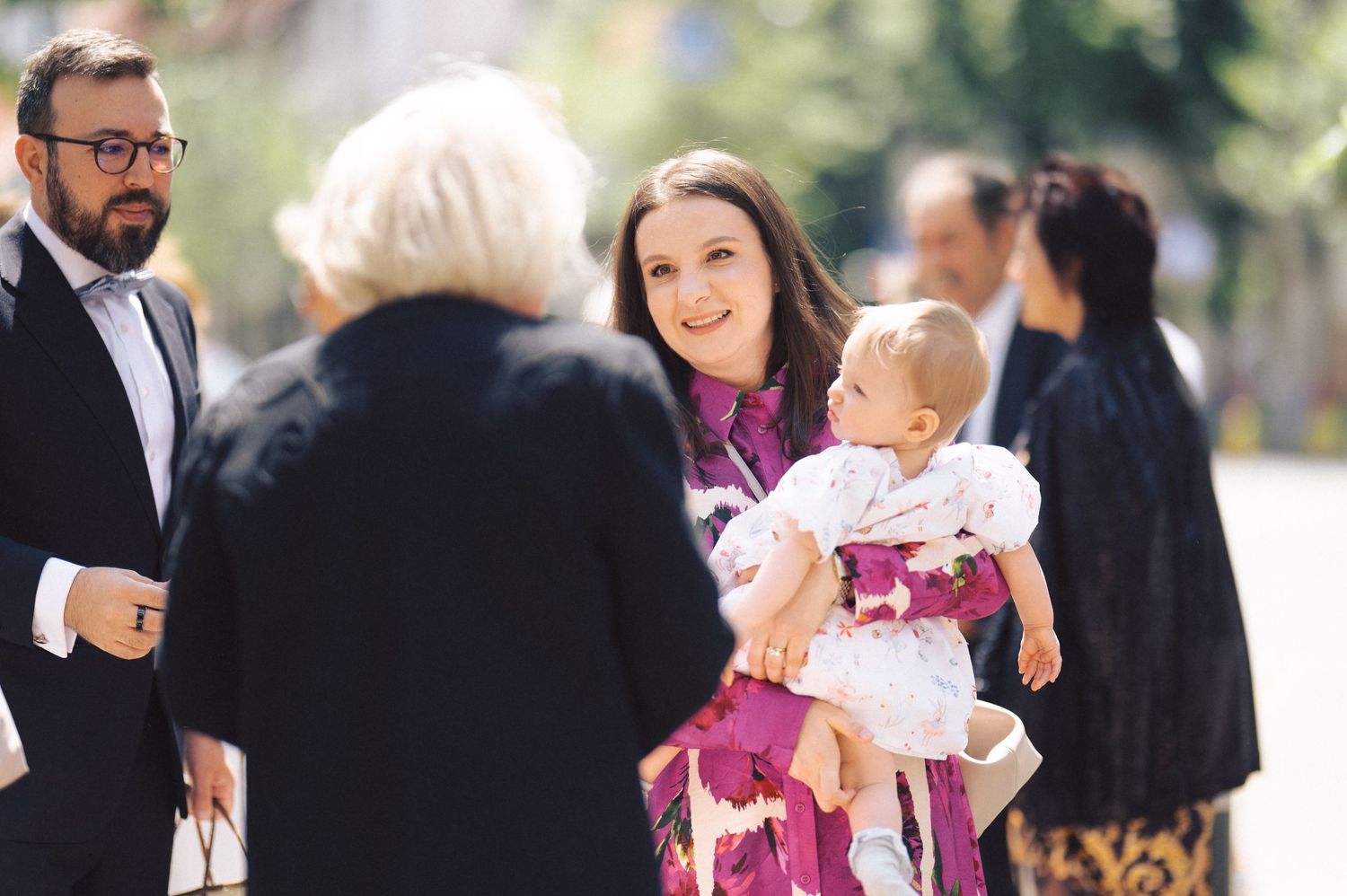 A person in a purple floral dress holds a baby in white at an outdoor social gathering.