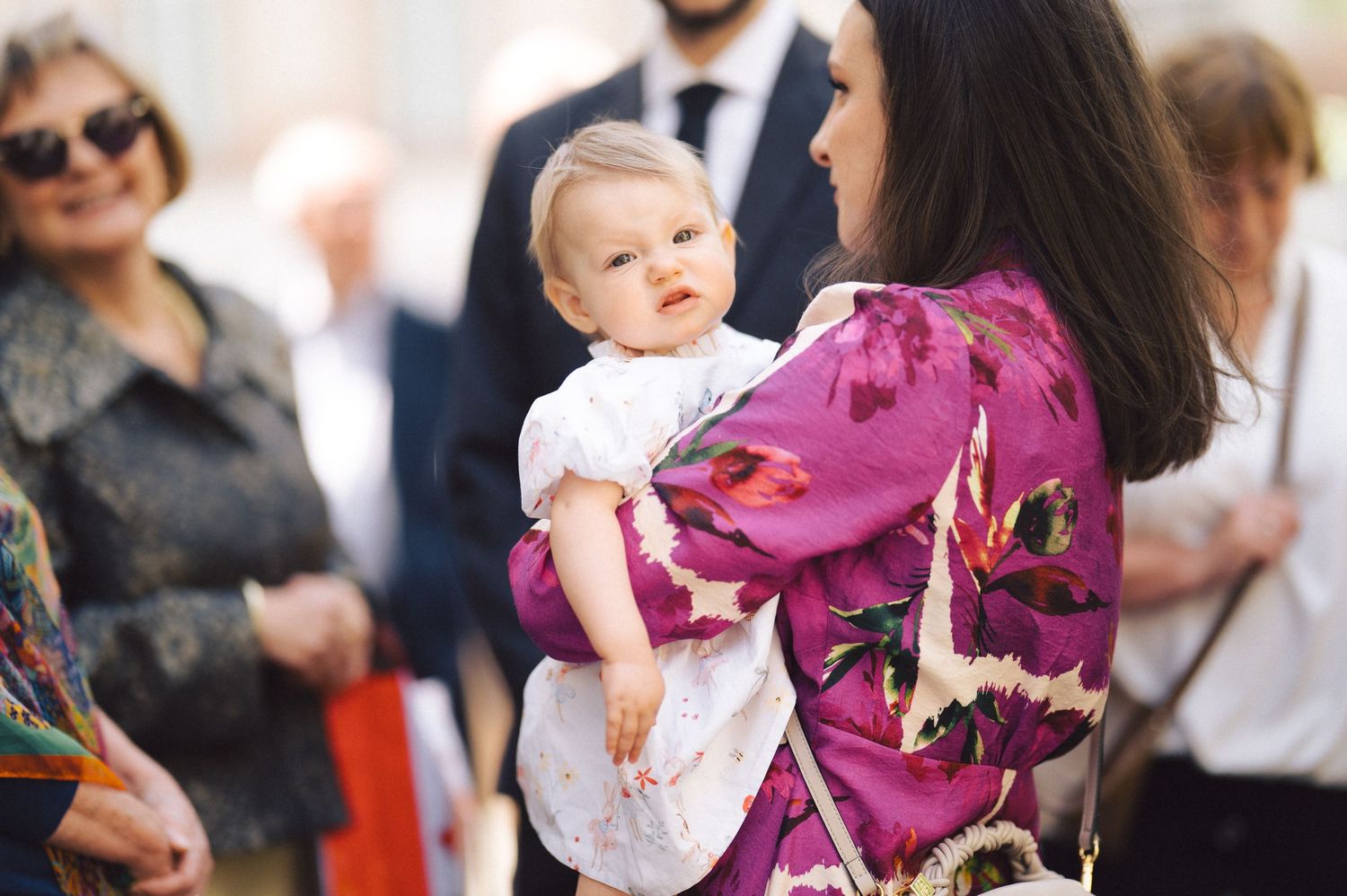 A person wearing a pink floral dress holds a small baby dressed in white at an event.