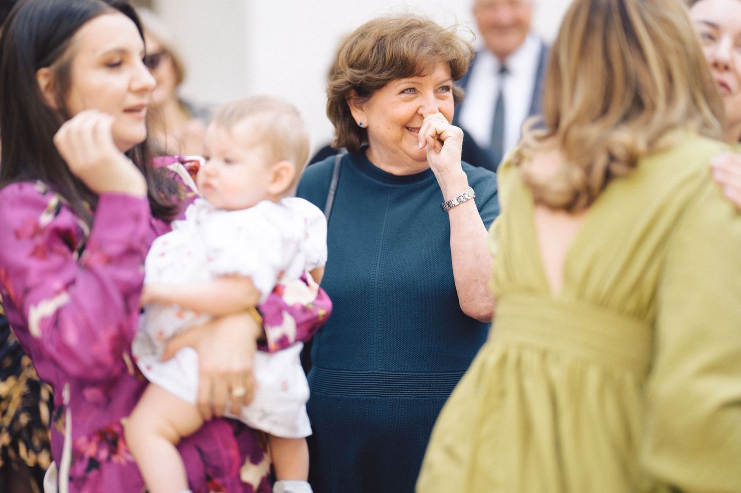 People gather in conversation at a social event with a baby while a woman in navy blue laughs joyfully.