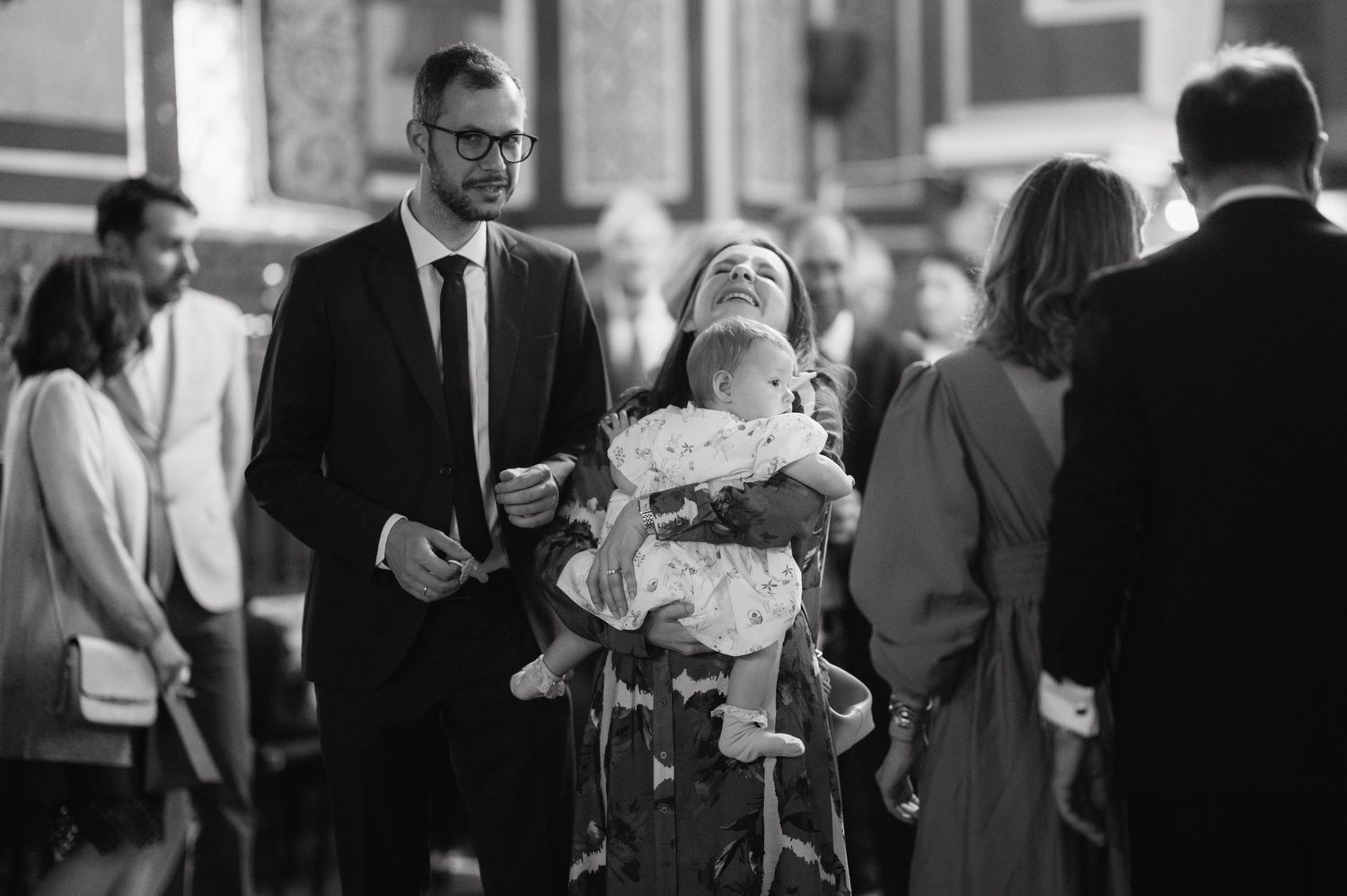 A black and white photograph shows a baptism ceremony in a church with people gathered around a baptismal font.