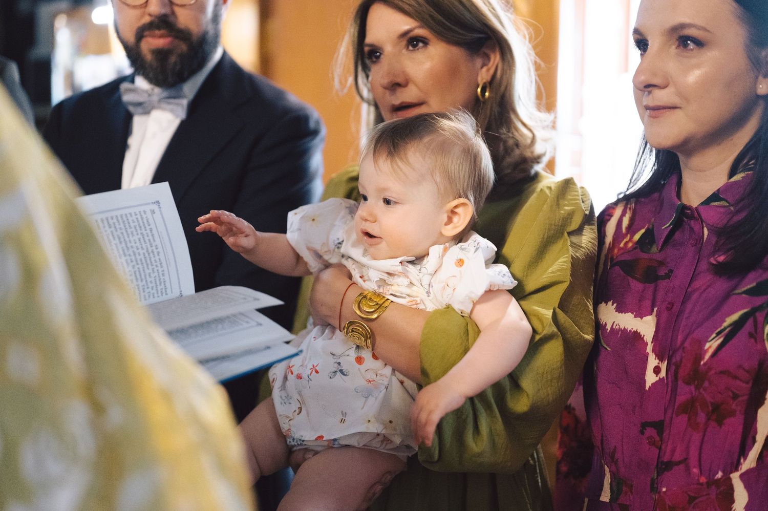 A baby wearing a white and yellow outfit celebrates their baptism ceremony surrounded by family members in church.