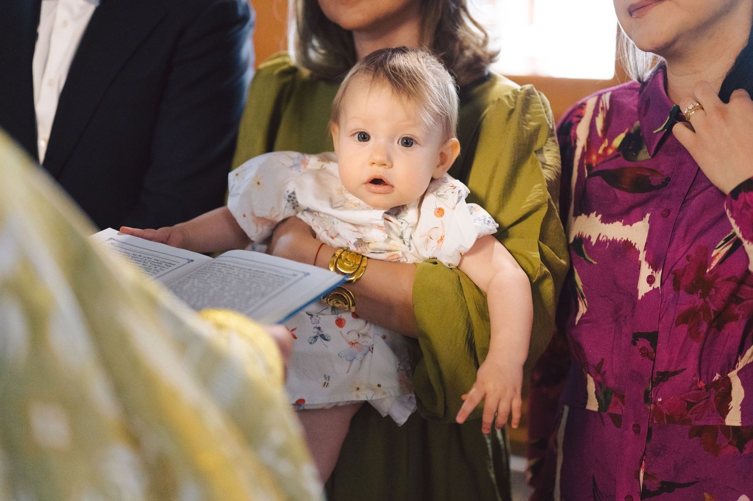 A small baby in a light-colored outfit sits between two adults wearing colorful clothing on a couch.