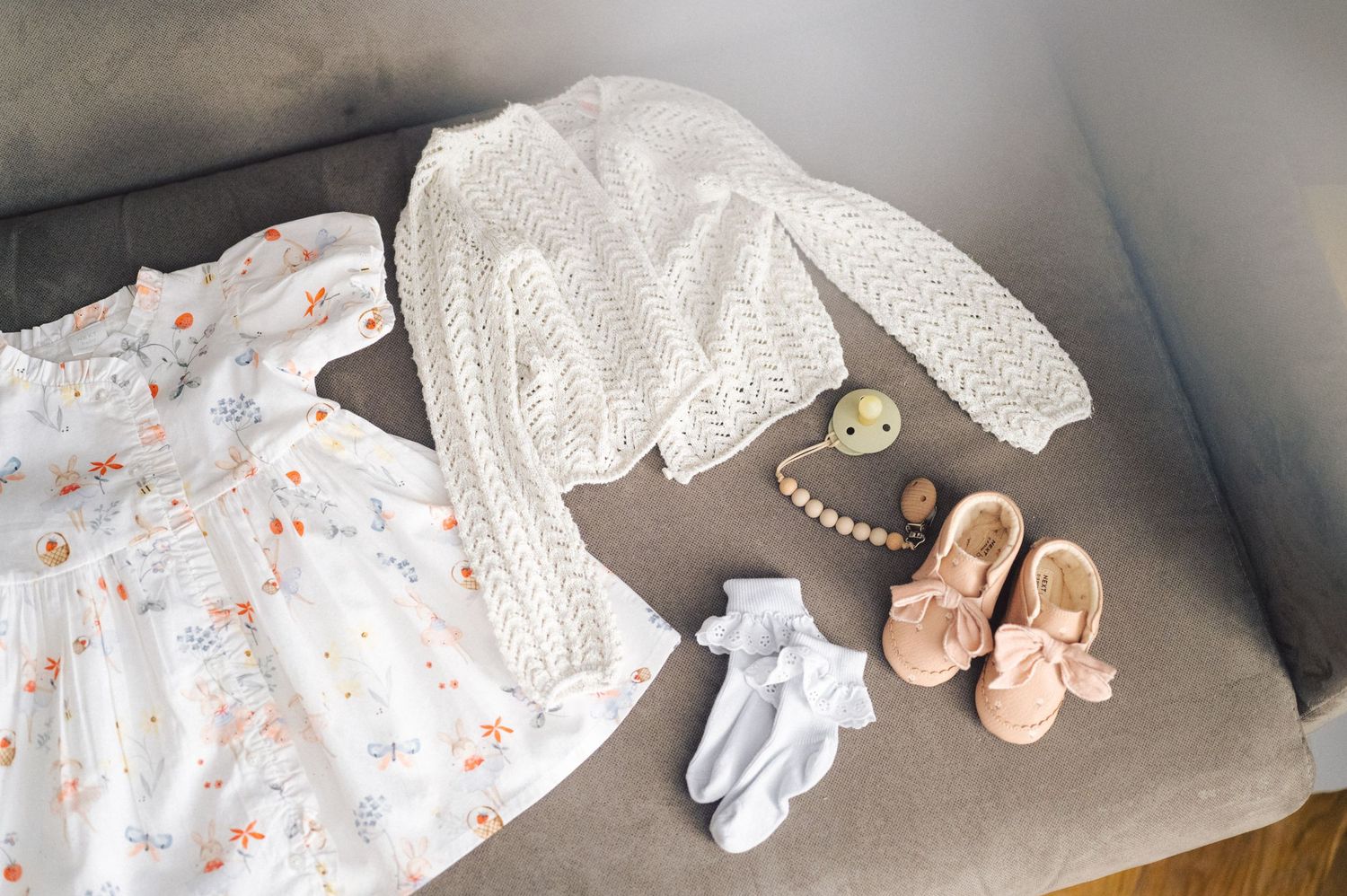 Baby clothing and accessories including a floral dress, white knit sweater, socks, and pink shoes laid out on grey surface.