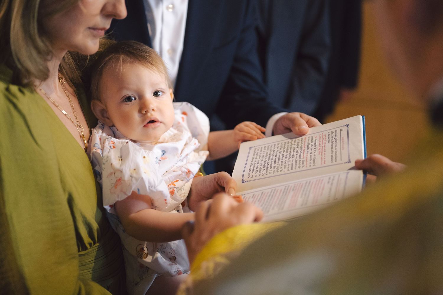 Baby in white clothing sits on guardian's lap during religious ceremony while reading from a text.